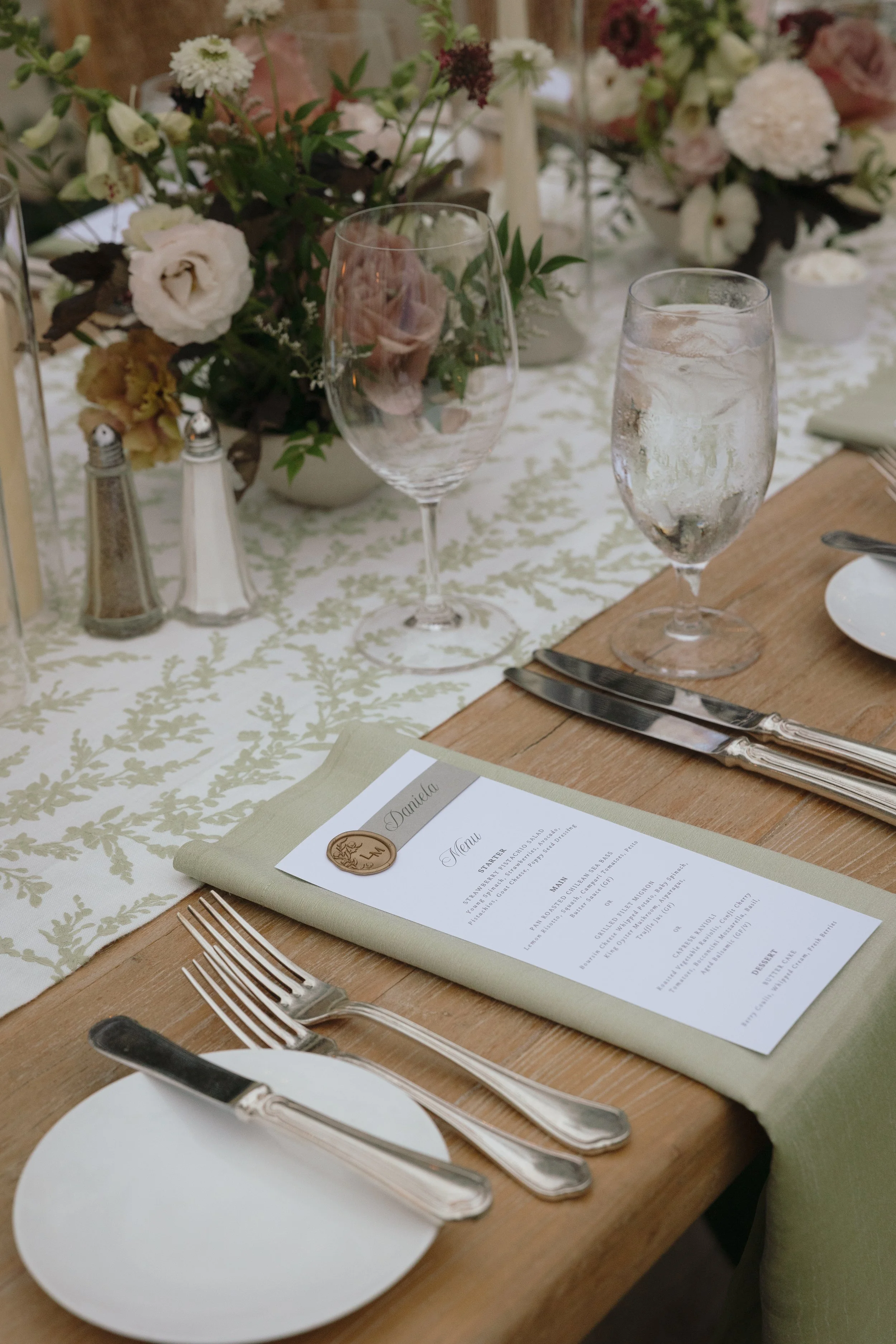 A formal dining table setup with a menu card, silverware, an empty white plate, and two glasses, one with water, on a wooden table with a floral table runner and a floral centerpiece in the background.