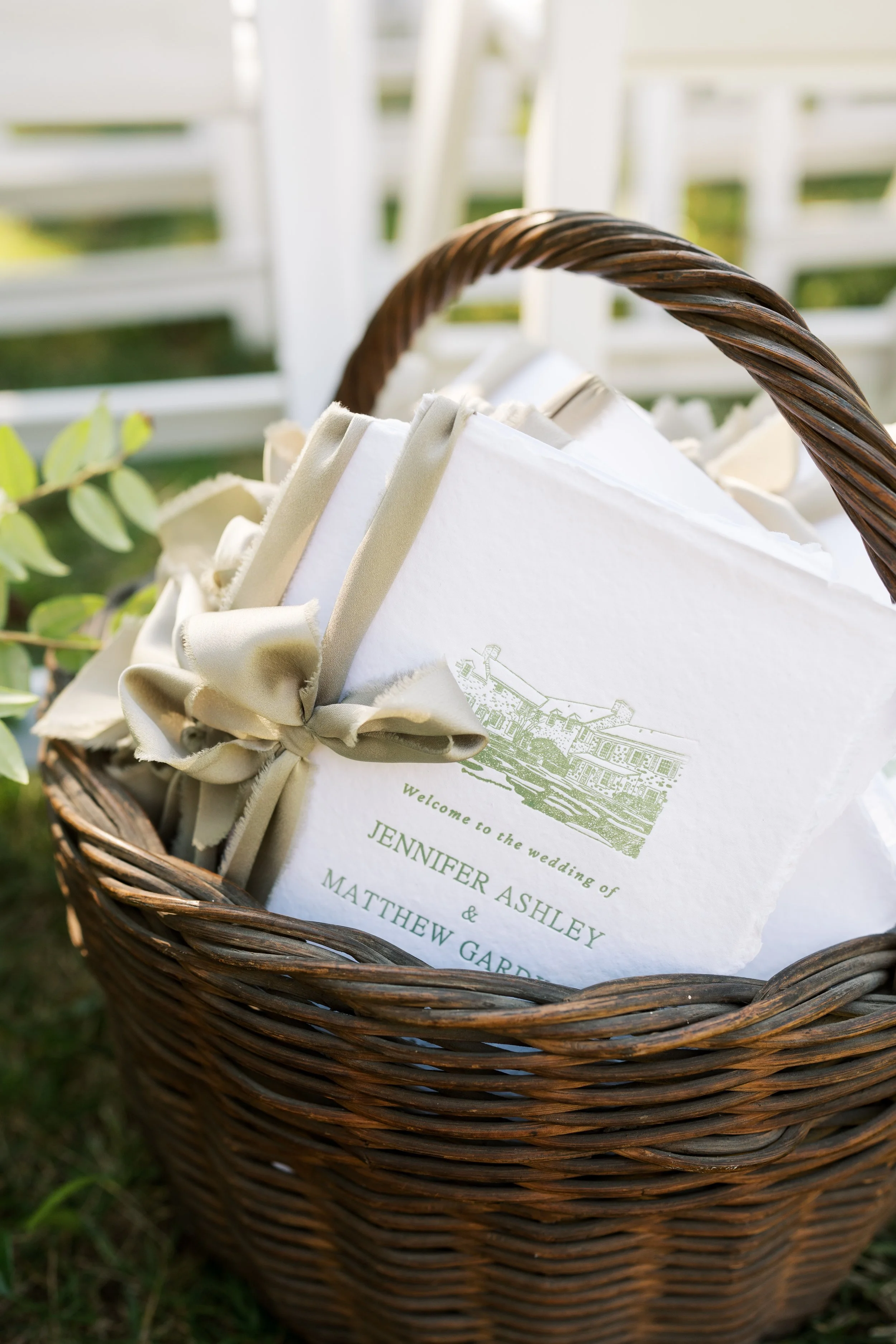 A wicker basket containing wedding programs with a beige ribbon, outdoors with greenery and white fencing in the background.