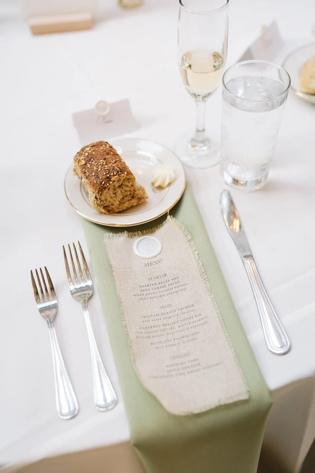 A table set for a meal with a bread roll, a menu, two glasses of water, a glass of white wine, and silverware.