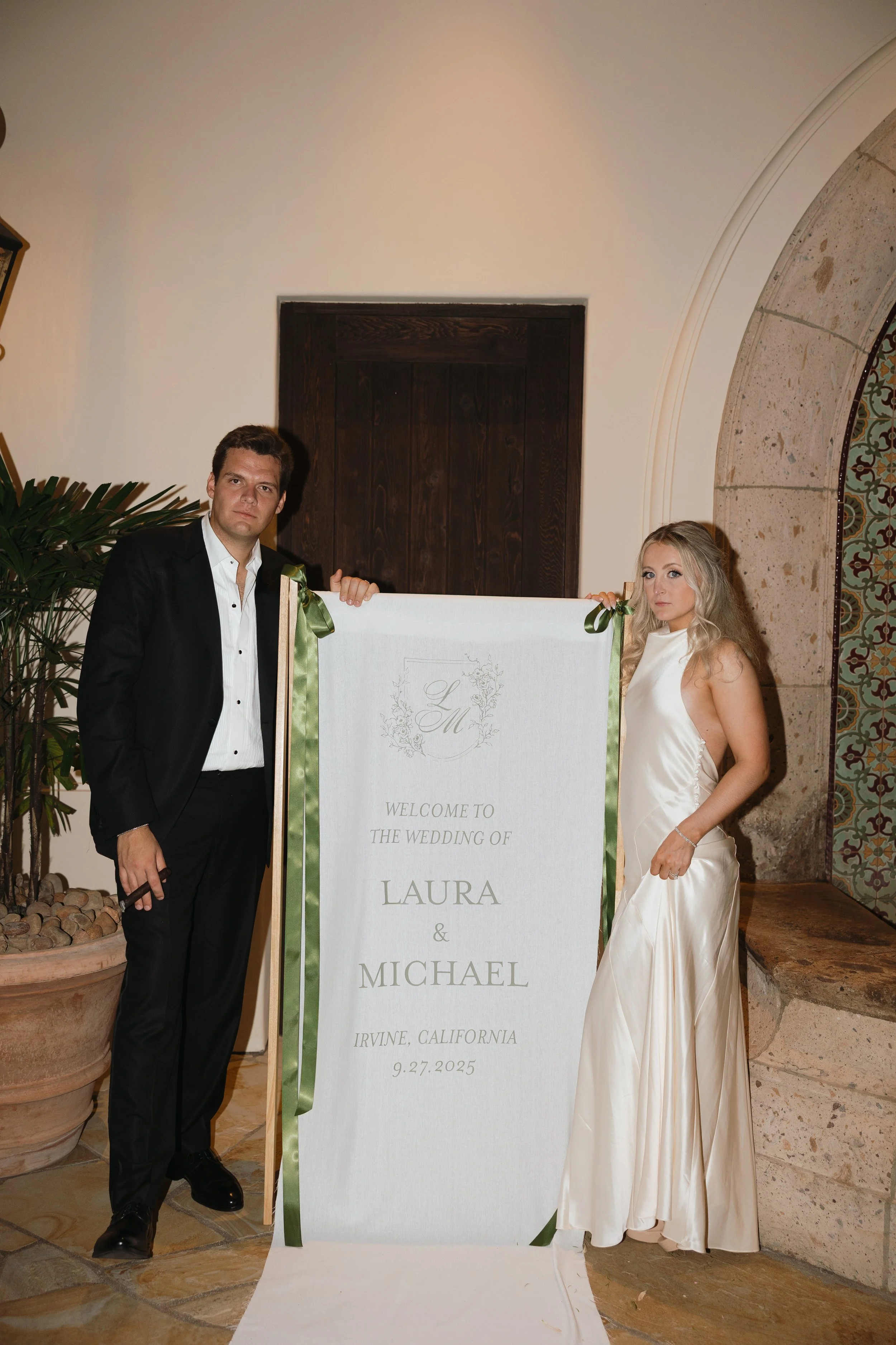 A man in a black suit and white shirt, and a woman in a white satin dress standing beside a wedding welcome sign for Laura and Michael. The sign indicates the wedding is in Irvine, California, on September 27, 2025.