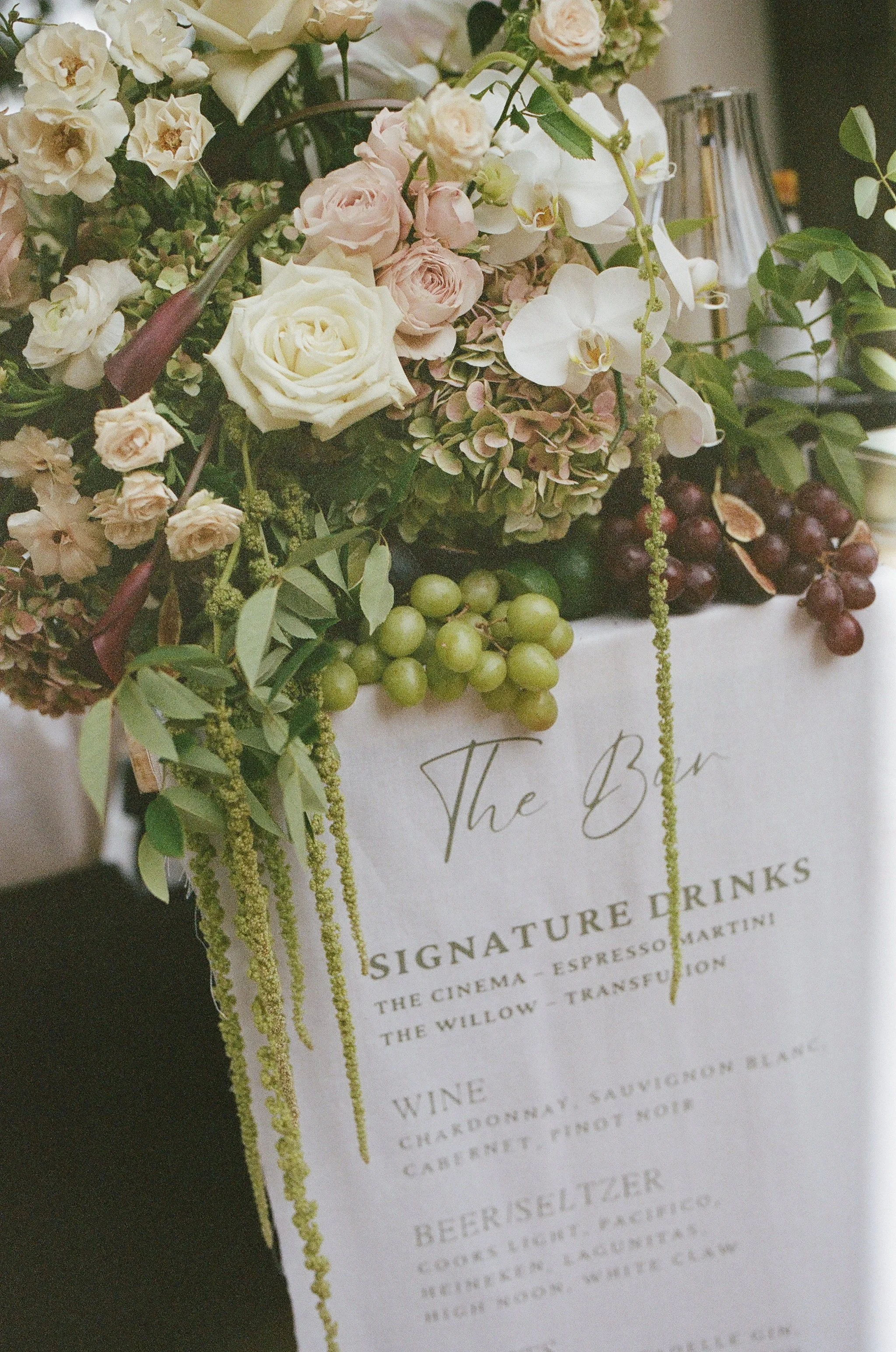 A floral arrangement with white roses, pink roses, white orchids, and greenery placed over a menu listing signature drinks.