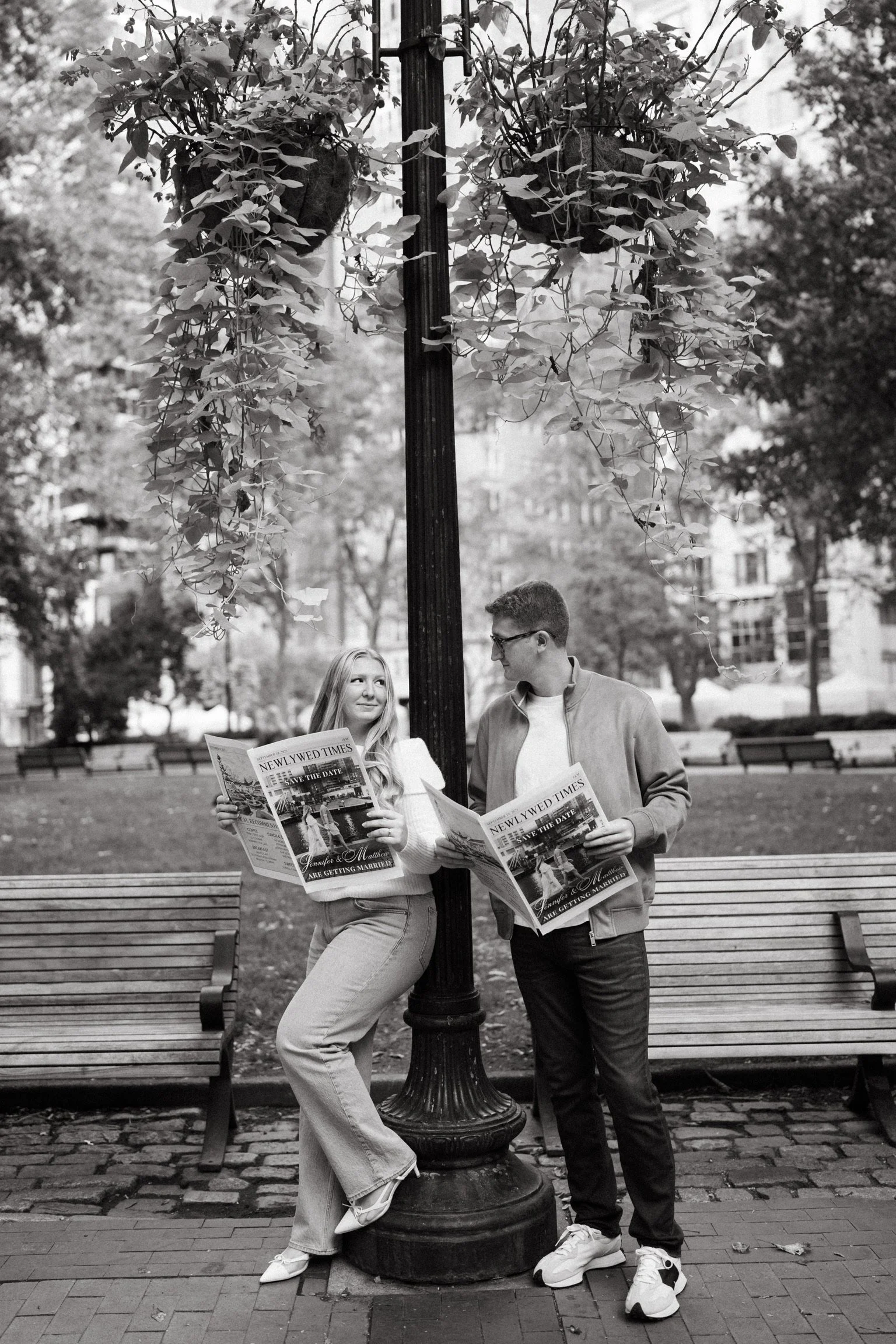 A black and white photo of a young man and woman standing on a park sidewalk, leaning against a lamppost wrapped with leafy vines, both reading newspapers.
