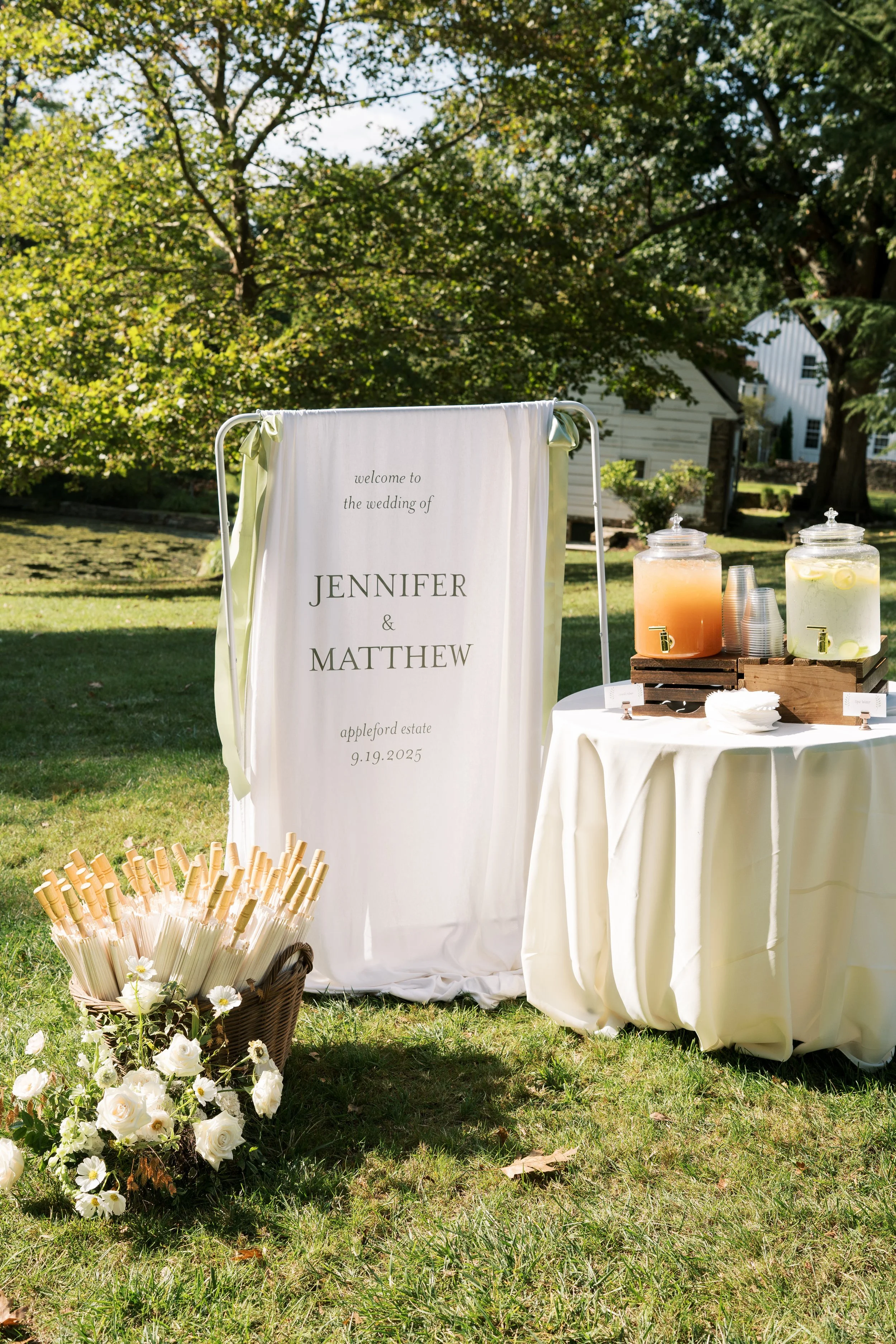 Wedding reception area outdoors with a welcome sign for Jennifer and Matthew's wedding on September 19, 2025, at Appleford Estate, surrounded by greenery, flowers, and lemonade dispensers.