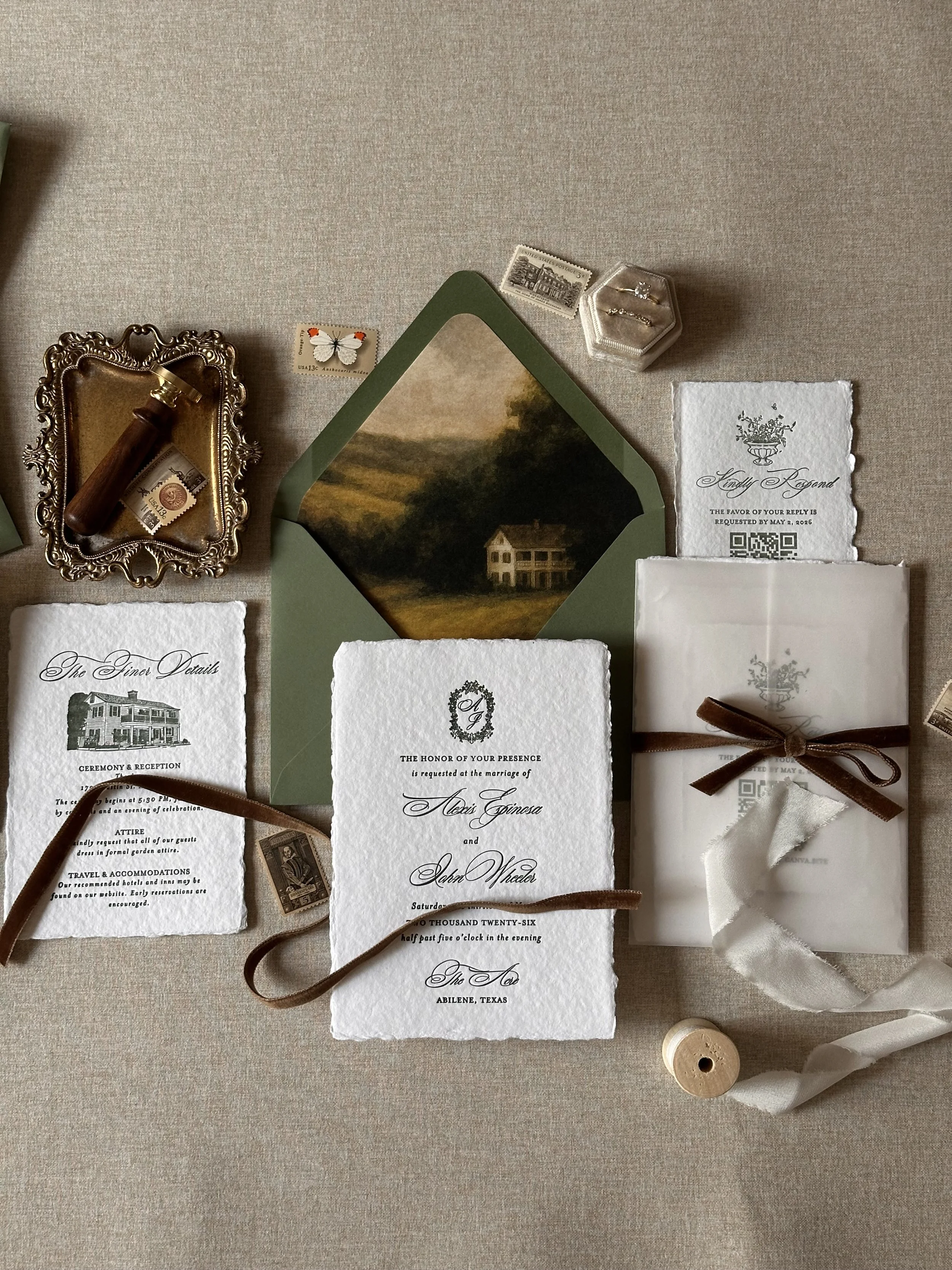 Wedding invitation suite includes ornate white and textured paper cards, a green envelope with a landscape painting, ribbon, postage stamps, and vintage jewelry, all arranged on a light fabric surface.
