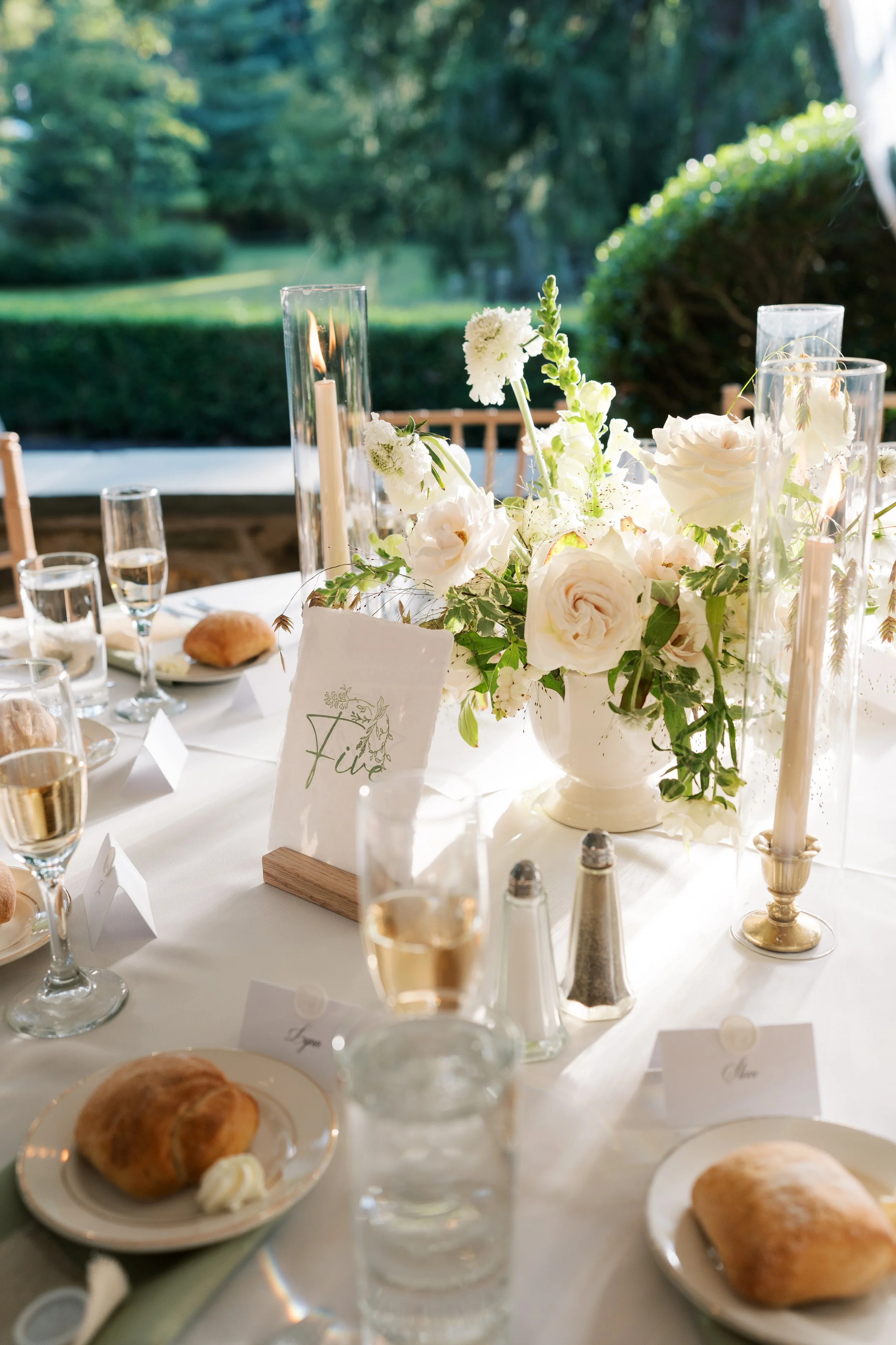Table set for a formal event outdoors with white flowers, candles, champagne, and bread rolls.