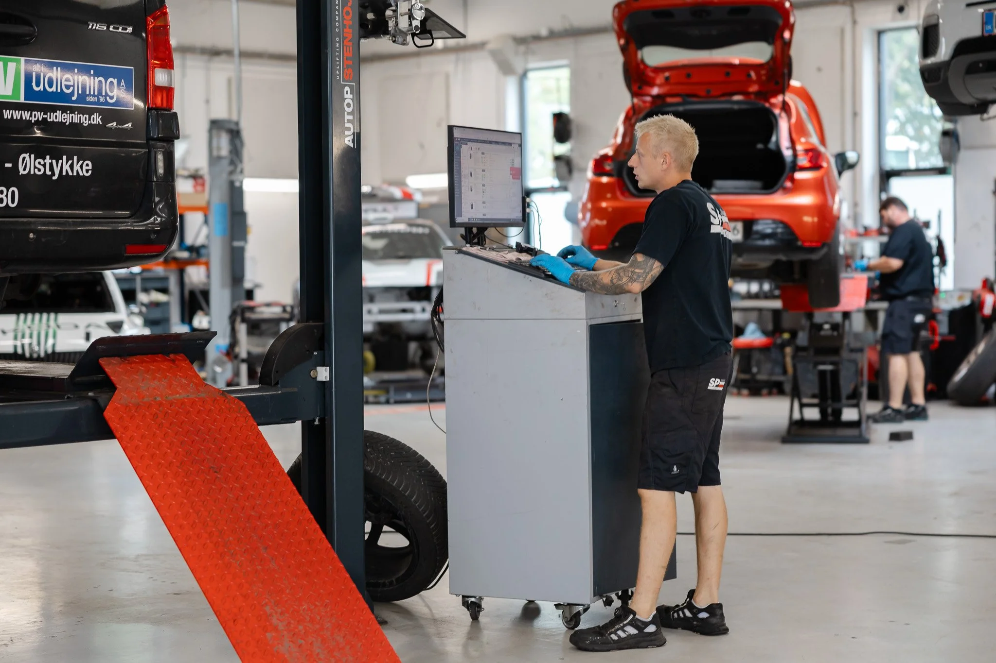 En mand med blond hår og tatoveringer på armen, iført sort t-shirt og shorts, arbejder i et autoværksted og ser på en computerskærm. Bag ham er en orange bil med åbent bagklap og flere bildele, og der er en anden person i baggrunden, der også arbejde