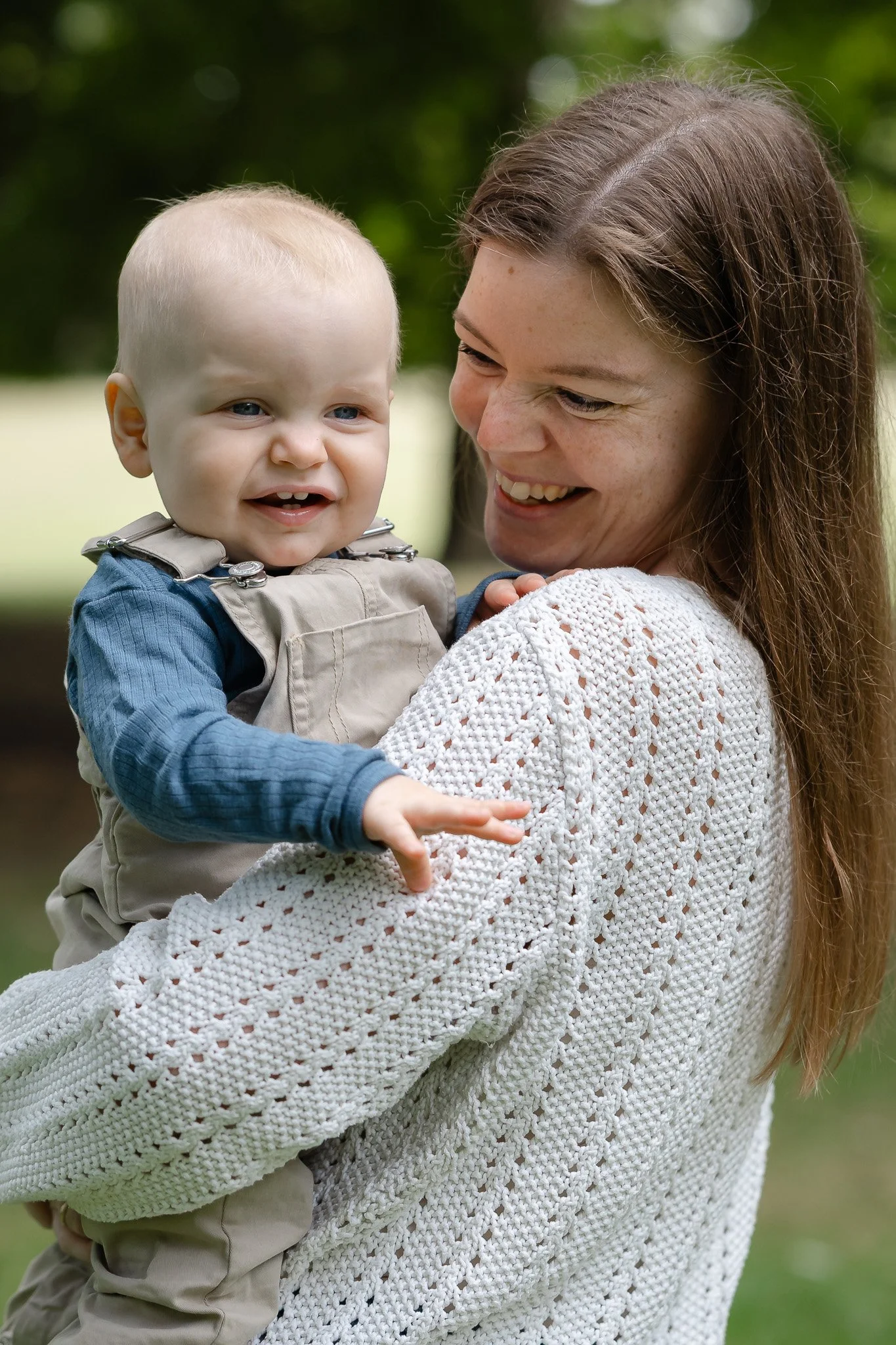 Mor holder sin smilende baby i armene under en naturlig familiefotografering udendørs.