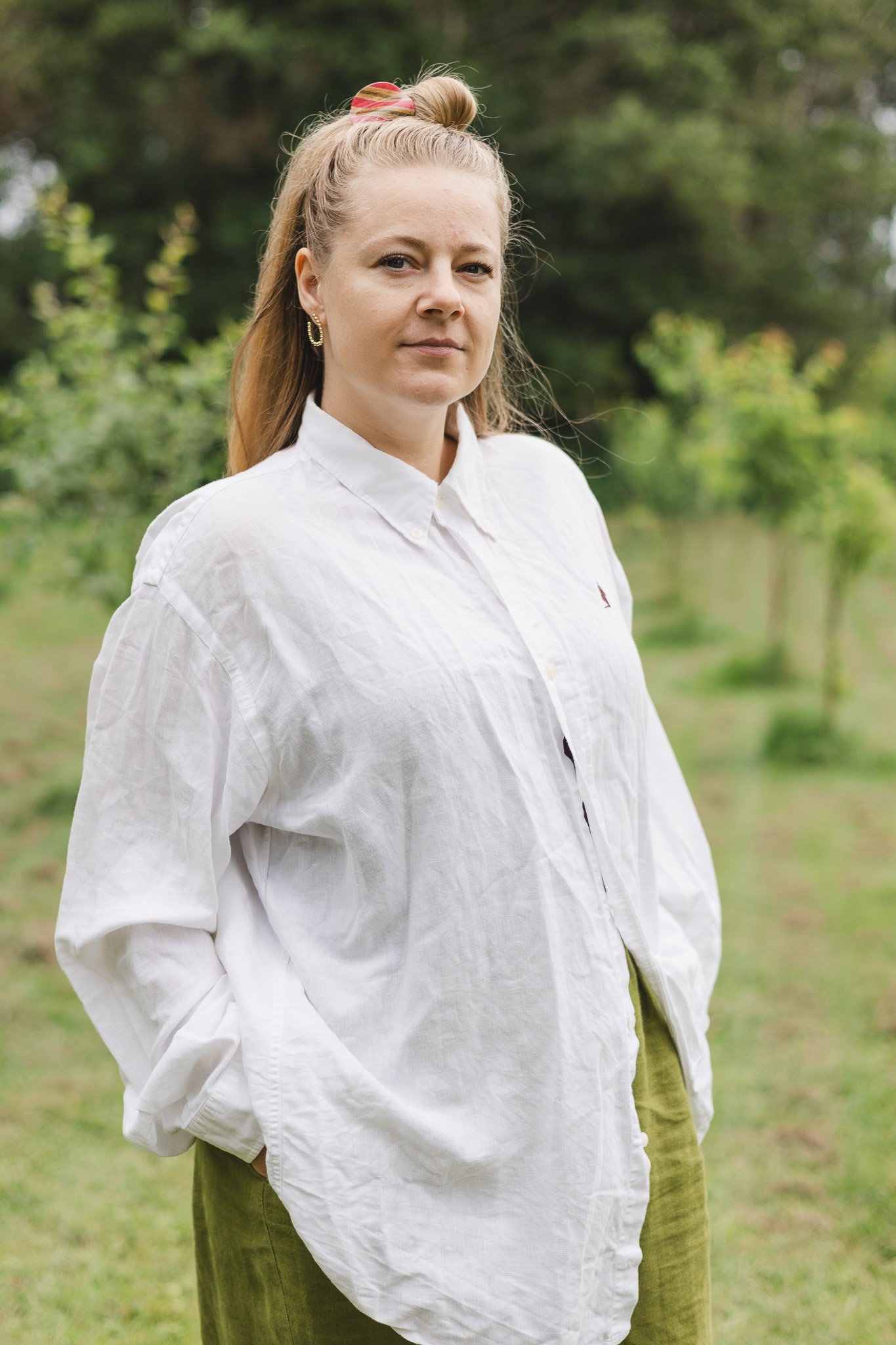Et portræt af en ung kvinde med langt blond hår i en knold, iført en hvid skjorte og grønne bukser, stående udendørs med grønne træer i baggrunden.