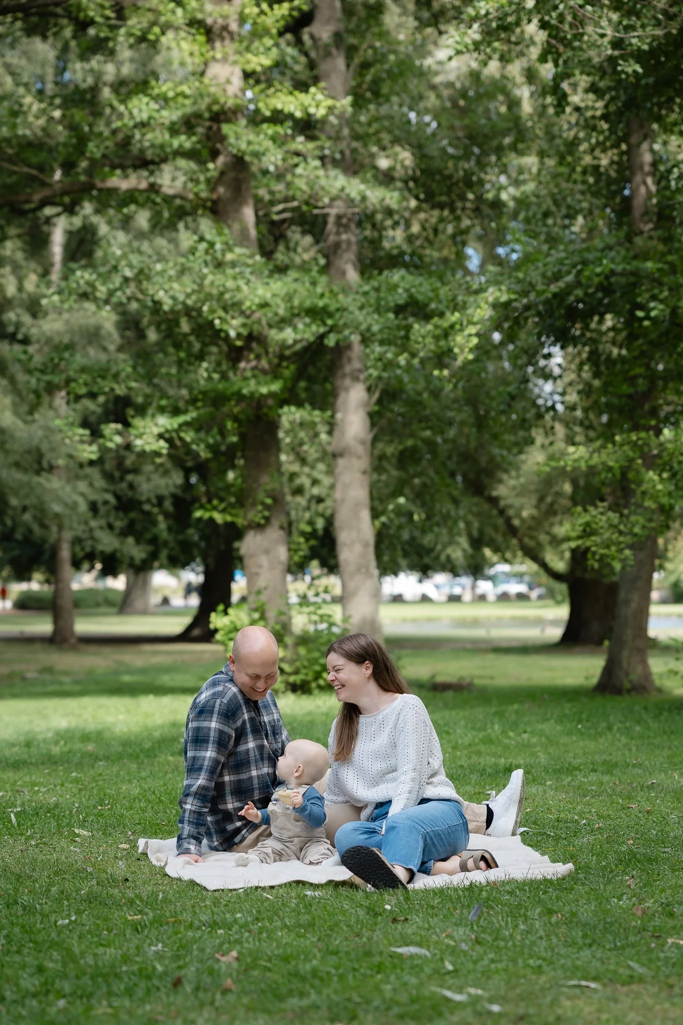 Familiefotografering i parken med mor, far og deres baby siddende sammen på et tæppe i det grønne græs.