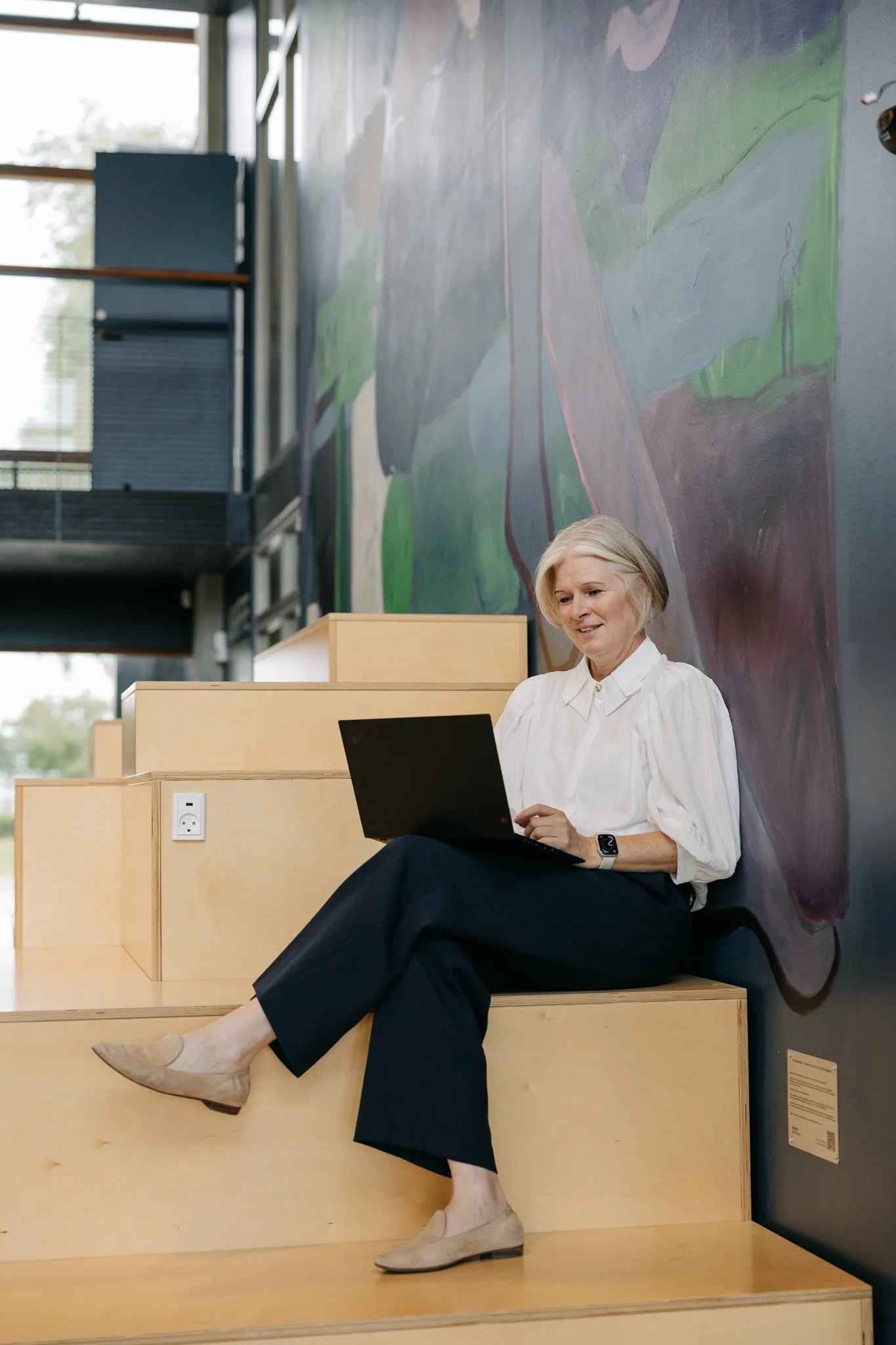 Kvindelig person sidder på trappetrin med ben krydset, bruger en bærbar computer, iført hvid skjorte, sorte bukser, beige sko og et armbåndsur, foran en væg med abstrakt kunst, inde i et moderne rum med store vinduer og træmøbler.