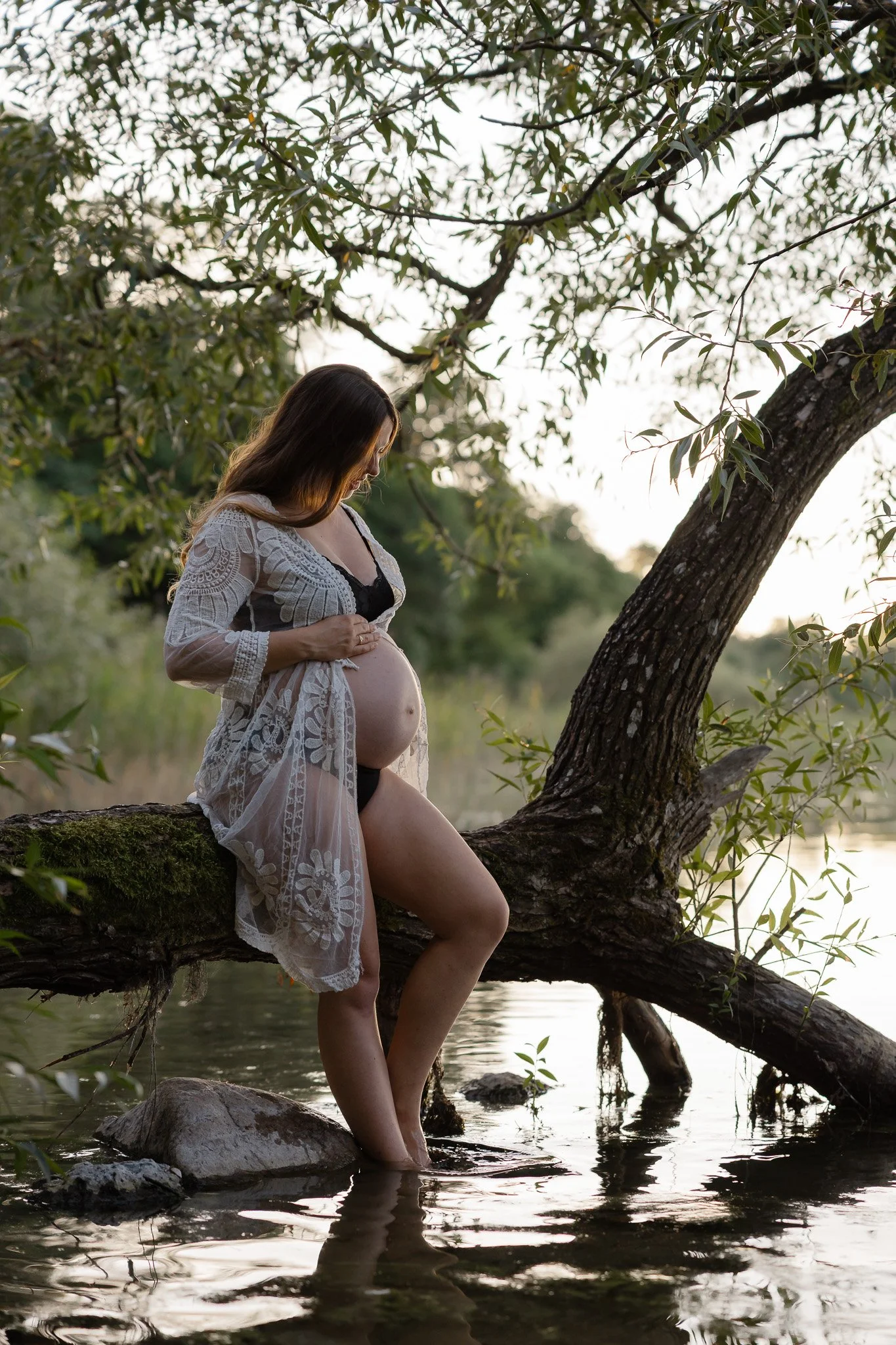 Gravid kvinde i en hvid blondekimono sidder på en træstamme omgivet af grønne træer under en udendørs graviditetsfotografering.