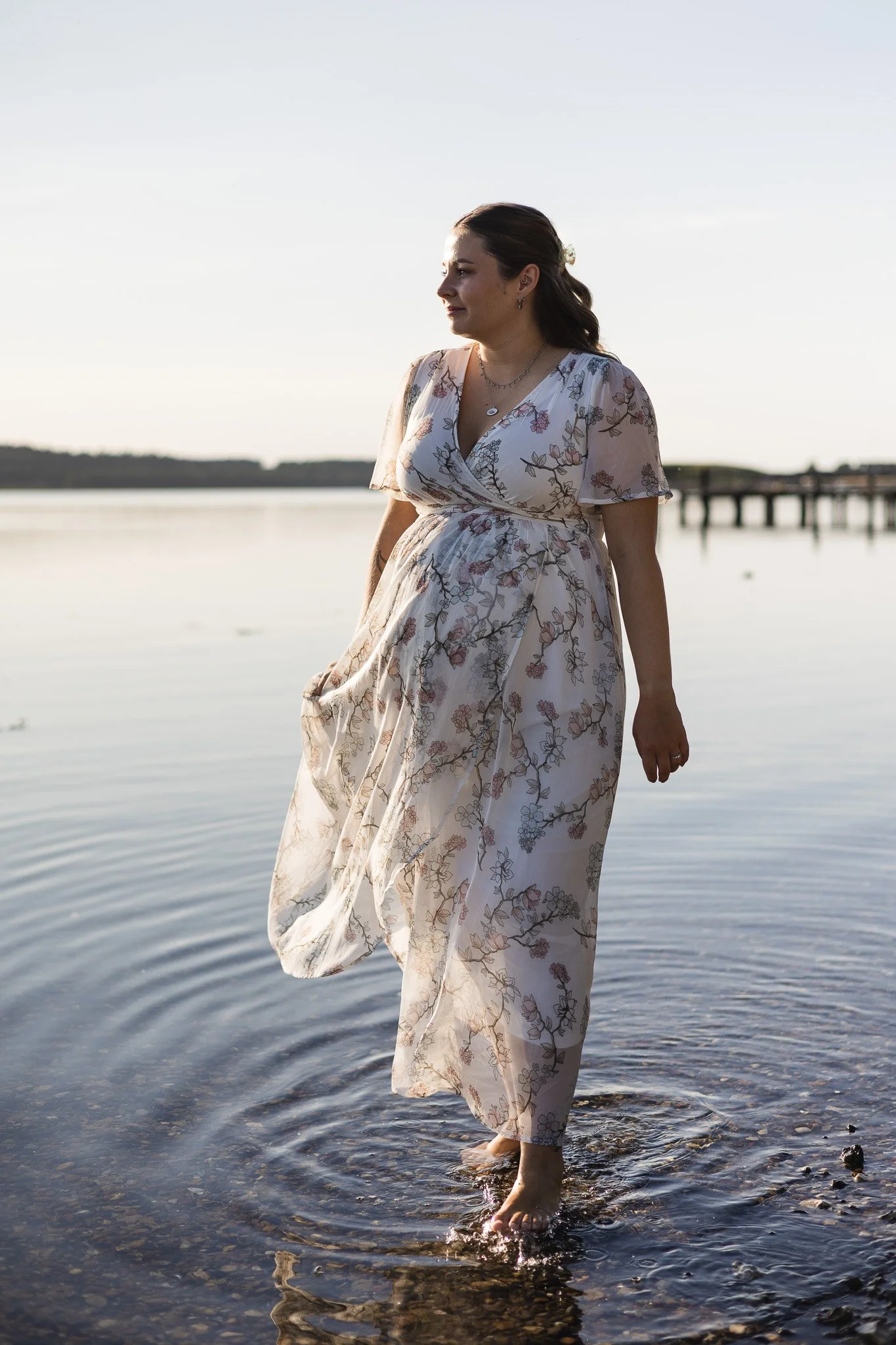 Gravid kvinde i en lang blomstret kjole står i det lave vand ved strandkanten i aftensolen.
