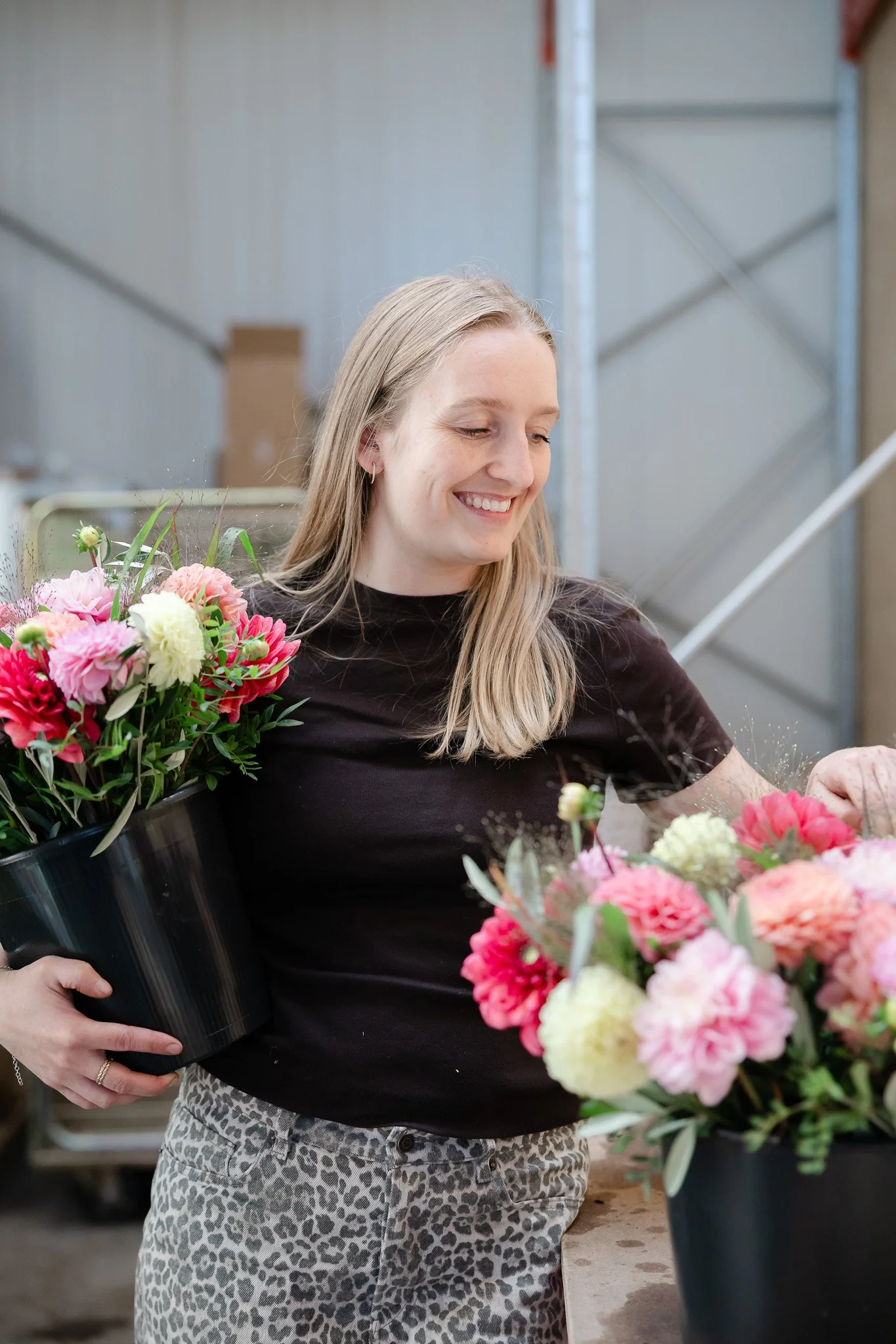 En smilende kvinde med blondt hår iført en sort T-shirt og leopardbukser, der holder en stor buket pink og hvid blomster, mens hun står ved et bord i et værksted eller butik.