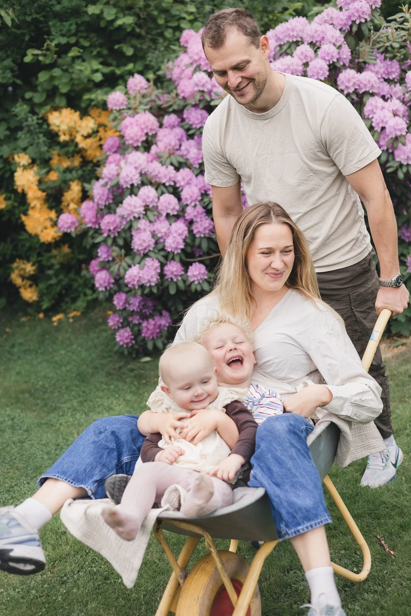 En familie leger udendørs foran blomstrende buske, hvor far skubber en barnevogn med tre børn, mens mor sidder i barnevognen og griner.