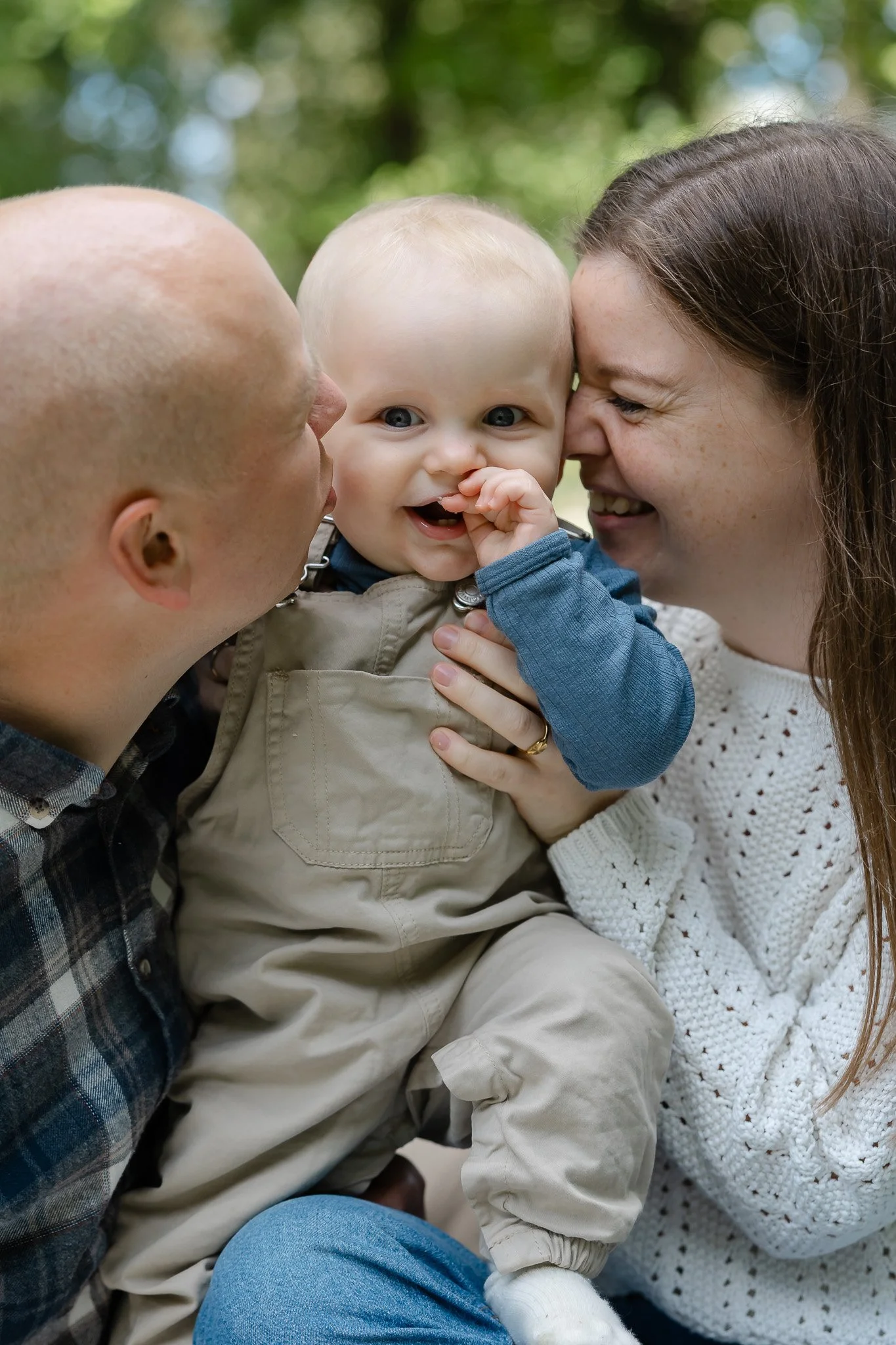 Mor og far kysser kærligt deres smilende baby under en naturlig familiefotografering udendørs