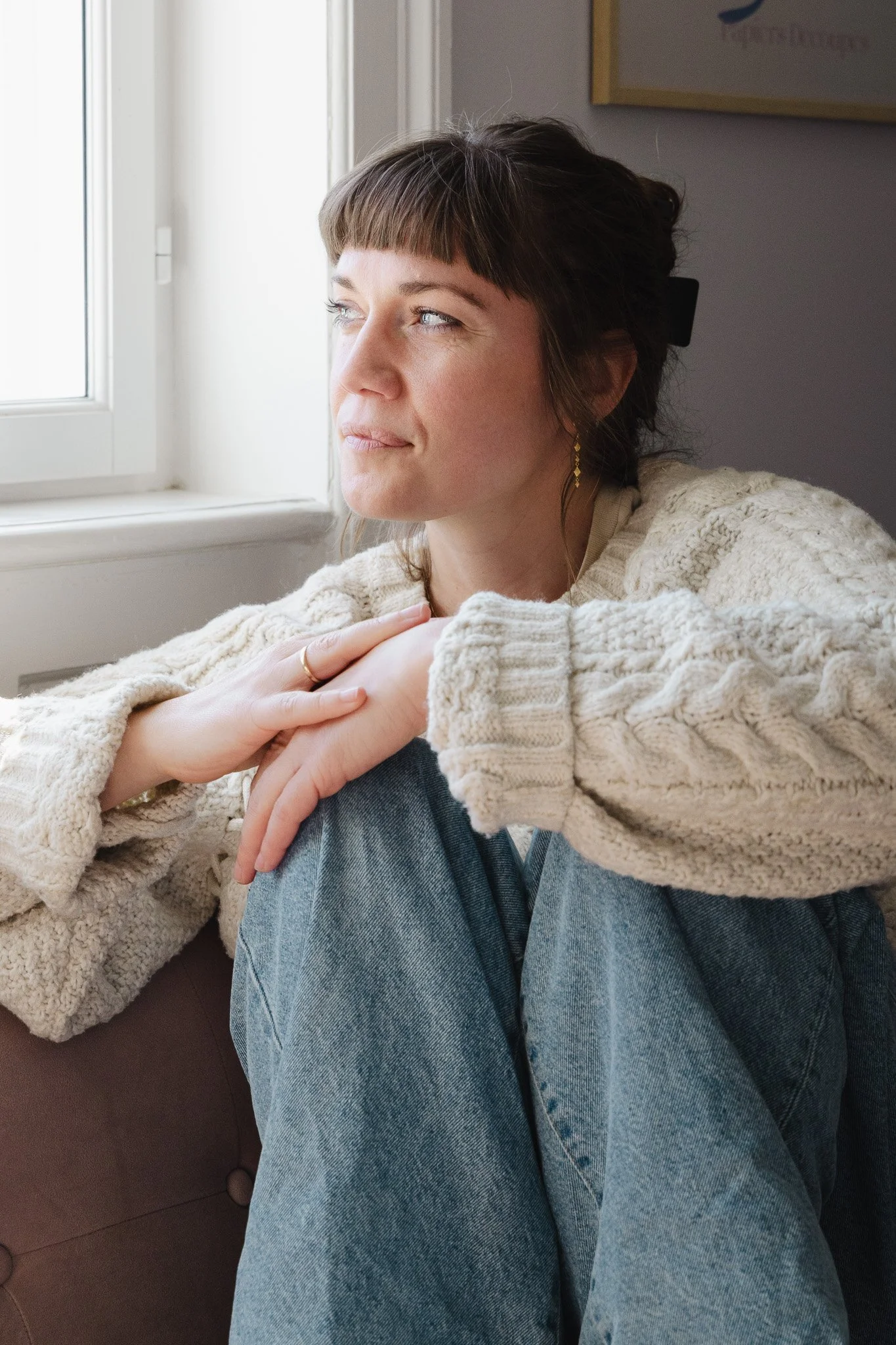 Kvinde sidder med knæene trukket op, hviler underholdt ved vinduet, iført en cremet sweater og jeans, i et rum med en malet væg.