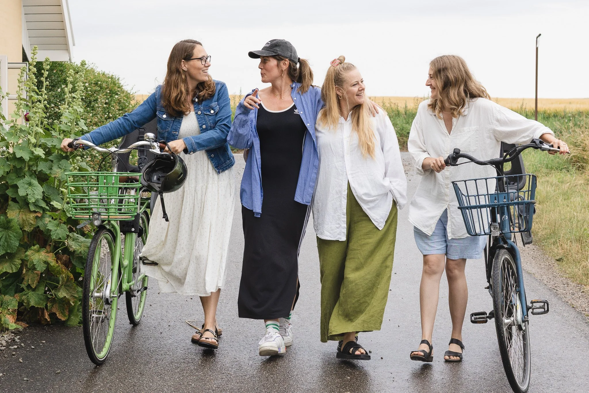 Fire kvinder går sammen på en vej med cykler, omgivet af grønt landbrug.