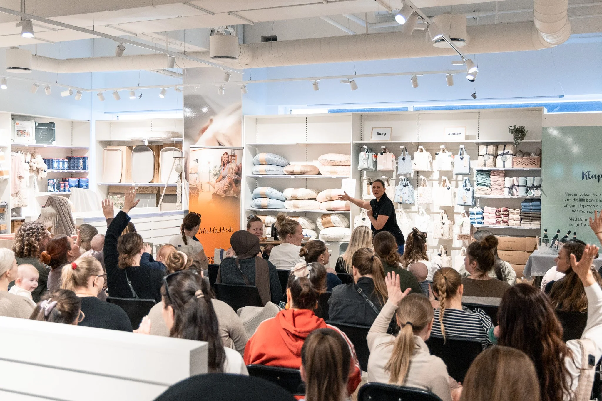 Et arrangement med mange voksne og børn, der sidder og lytter til en kvinde, der taler foran en butikshylde med tekstiler og babyprodukter i baggrunden.
