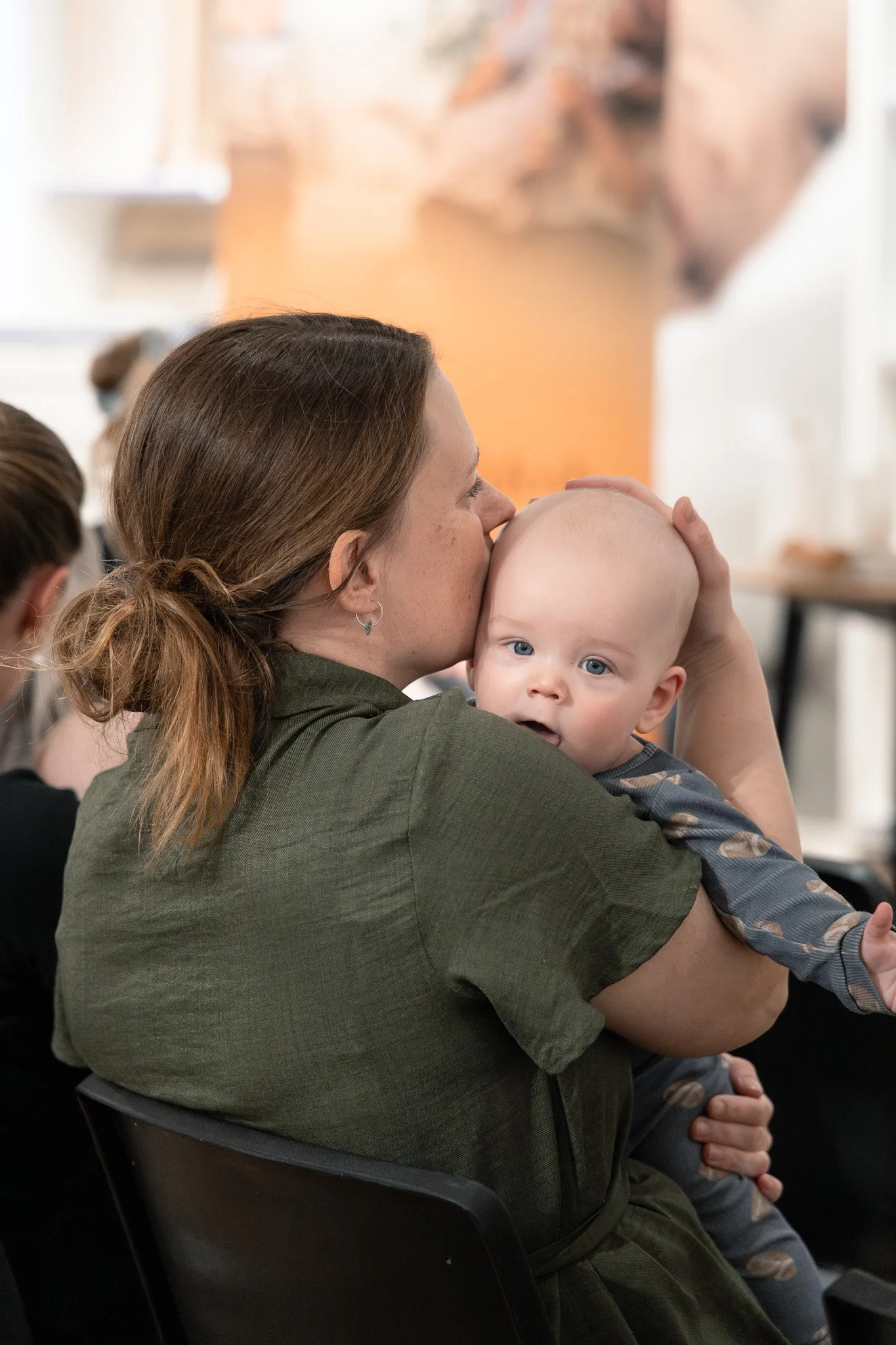 En kvinde med brunt hår i hestehale, der holder en baby med blå øjne og bart. Kvinden kysser babyens pande i et rørefuldt øjeblik.