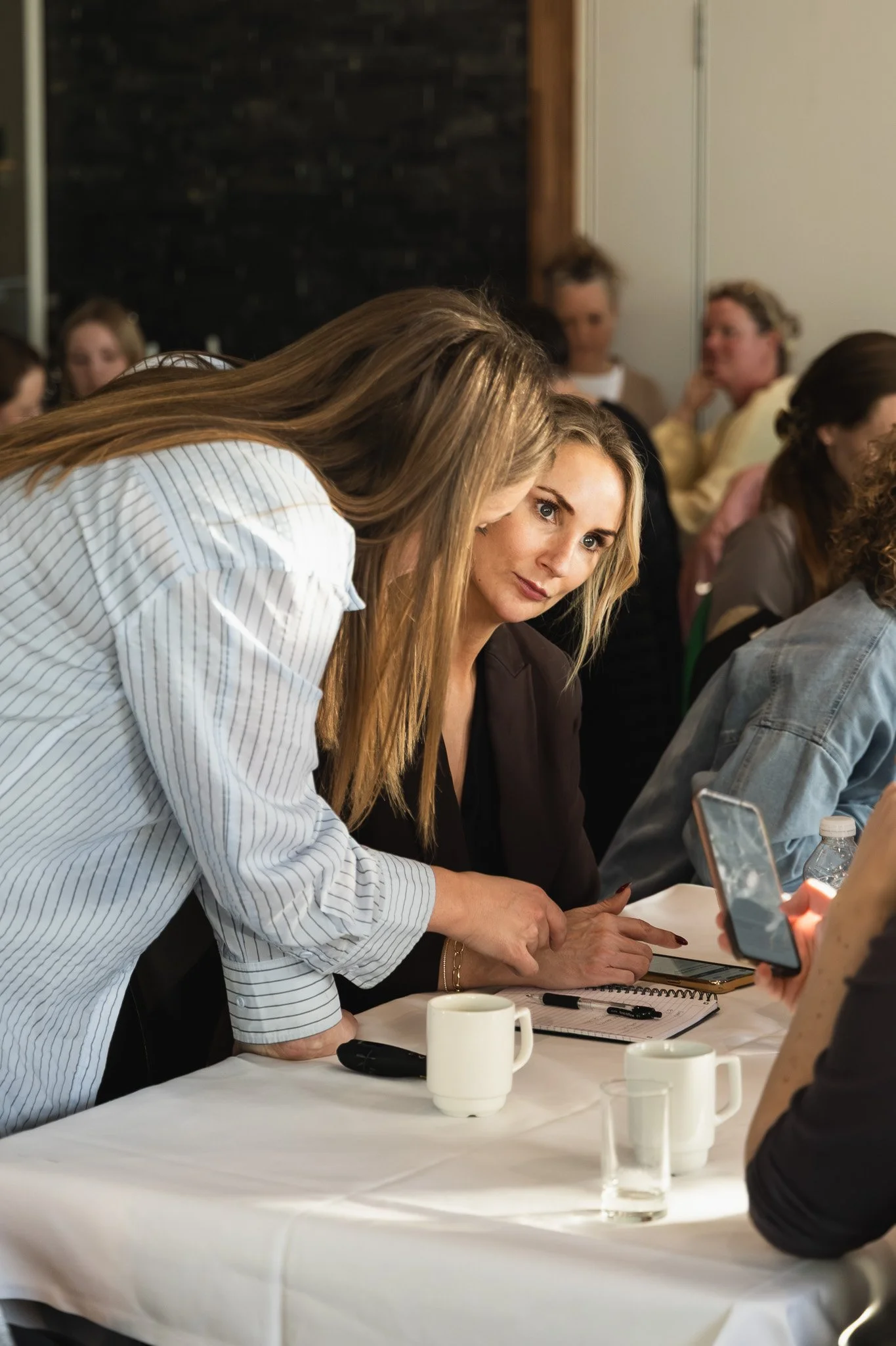 En gruppe mennesker sidder omkring et bord under en formel eller et møde. Nogle personer diskuterer, og en person står og skriver eller viser noget på sin telefon. Der er kaffekopper, notesbøger og skrevne noter på bordet.