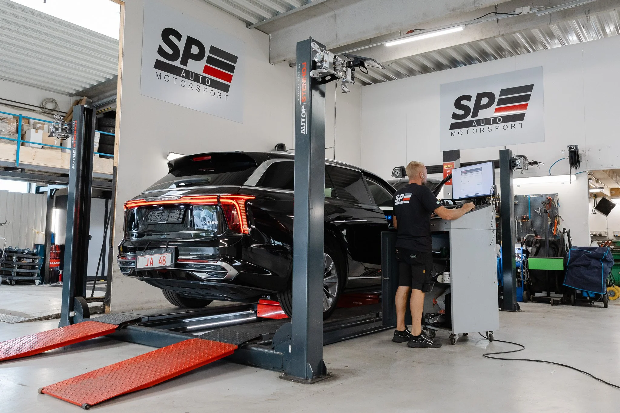En sort SUV bil befinder sig på en biltestbænk i et autoværksted, hvor en mand arbejder foran en computer. Der er tydelige skilte med logoet 'SP AUTO MOTORSPORT' på væggen.