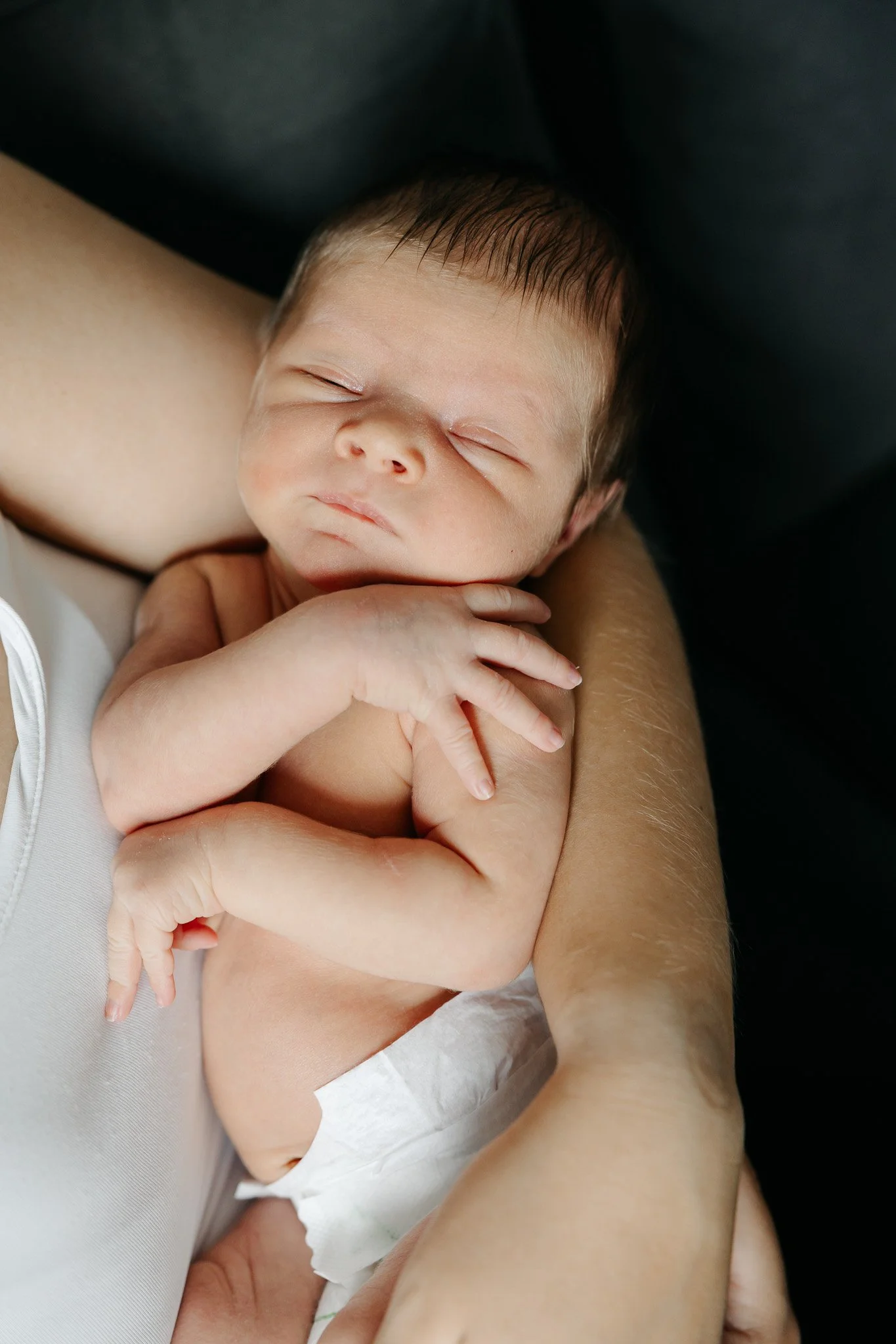 Nyfødt baby sover på mors arm med hænderne samlet foran kroppen.