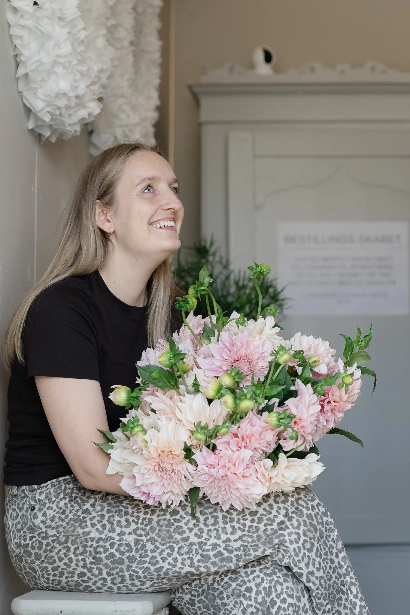 En glad kvinde holder en stor bunke af lyserøde og hvide blomster, sidder på en stol, med en beige leopardprint nederdel og sort T-shirt, med en beige væg i baggrunden.