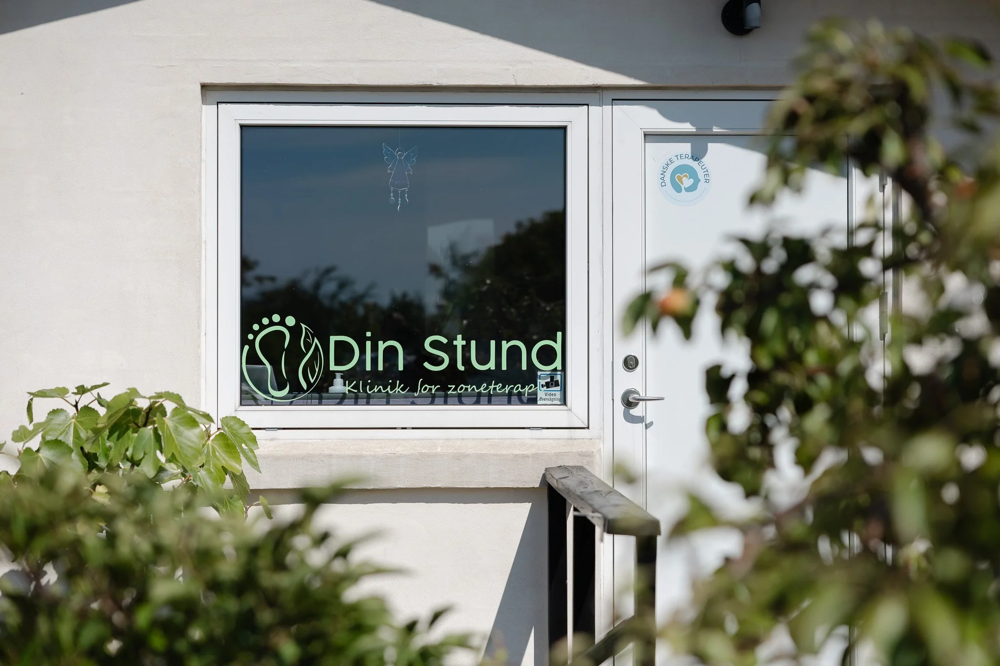Billedet viser indgangen til en klinik for zoneterapi, kaldet 'Din stund', med en bogstavelig facade og vindue med logo og dekorationer, omgivet af grønne blade.