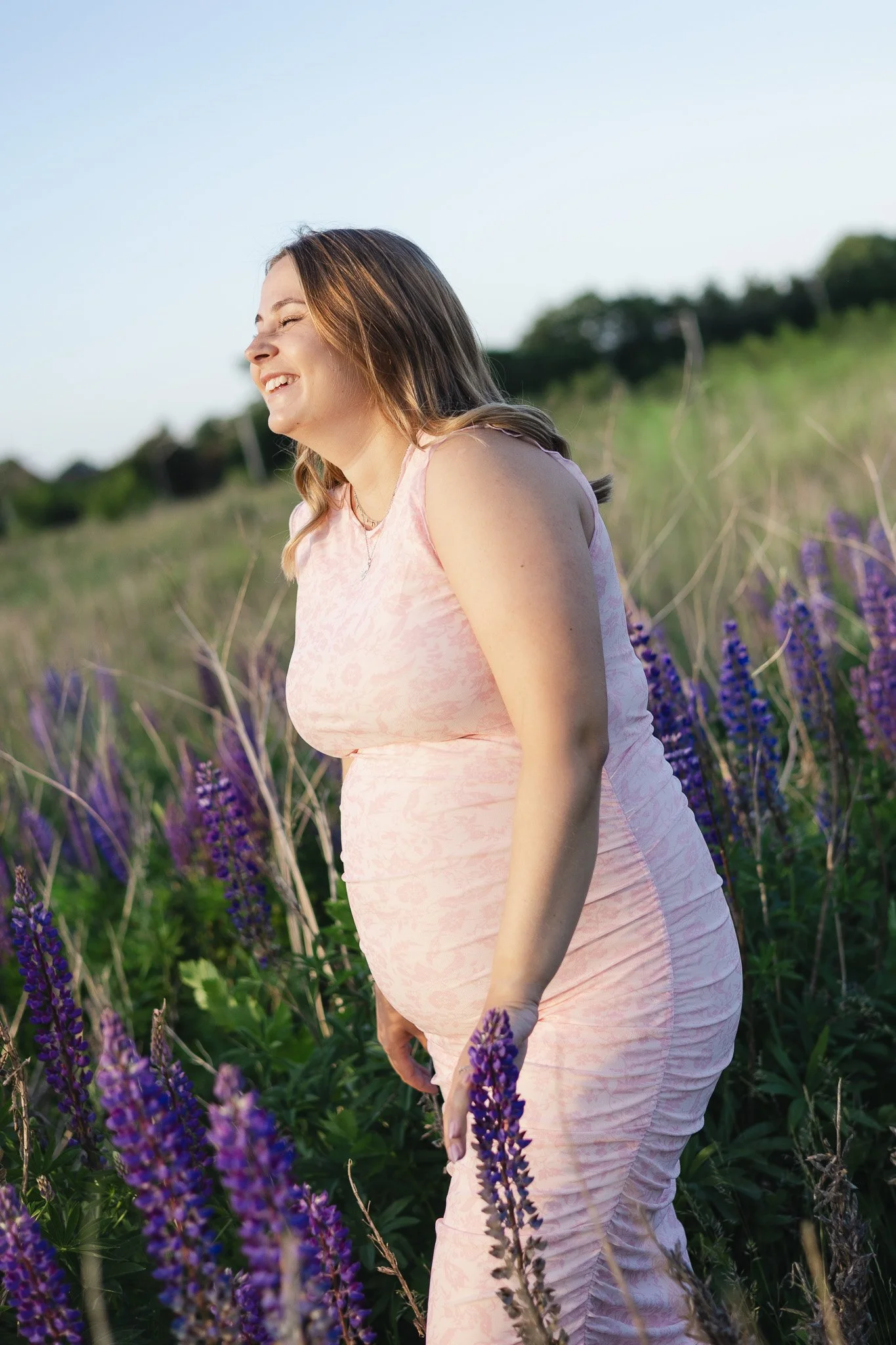 Gravid kvinde i en lyserød kjole omgivet af lilla blomster under en udendørs graviditetsfotografering.