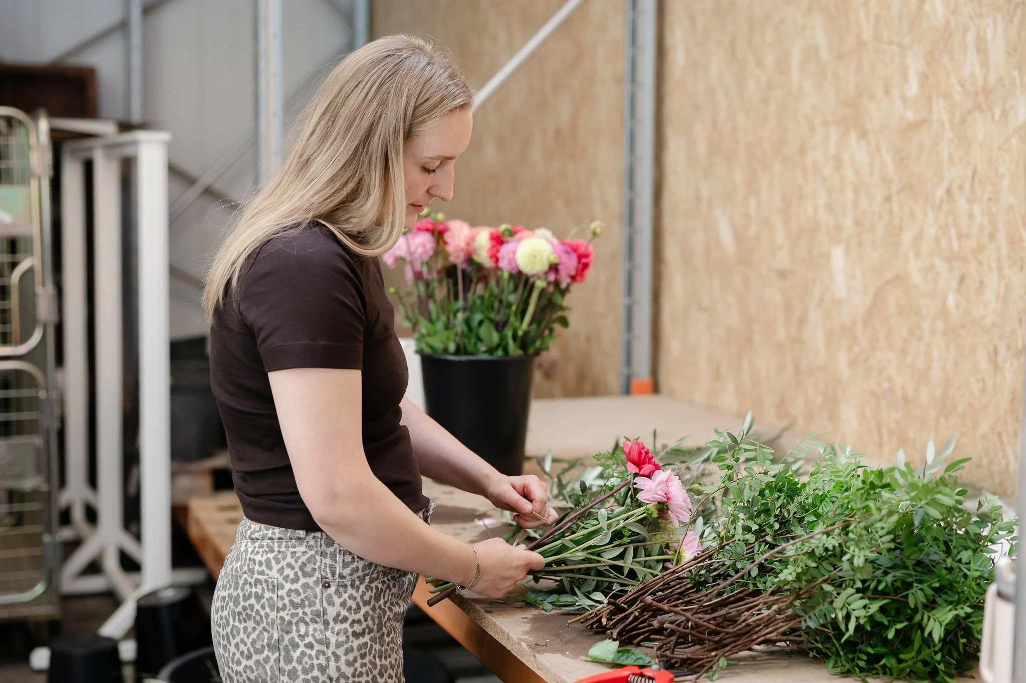 En kvinde arrangerer blomster i et værksted. Bag hende står en stor blomsterkum og en væg af trælæsning.