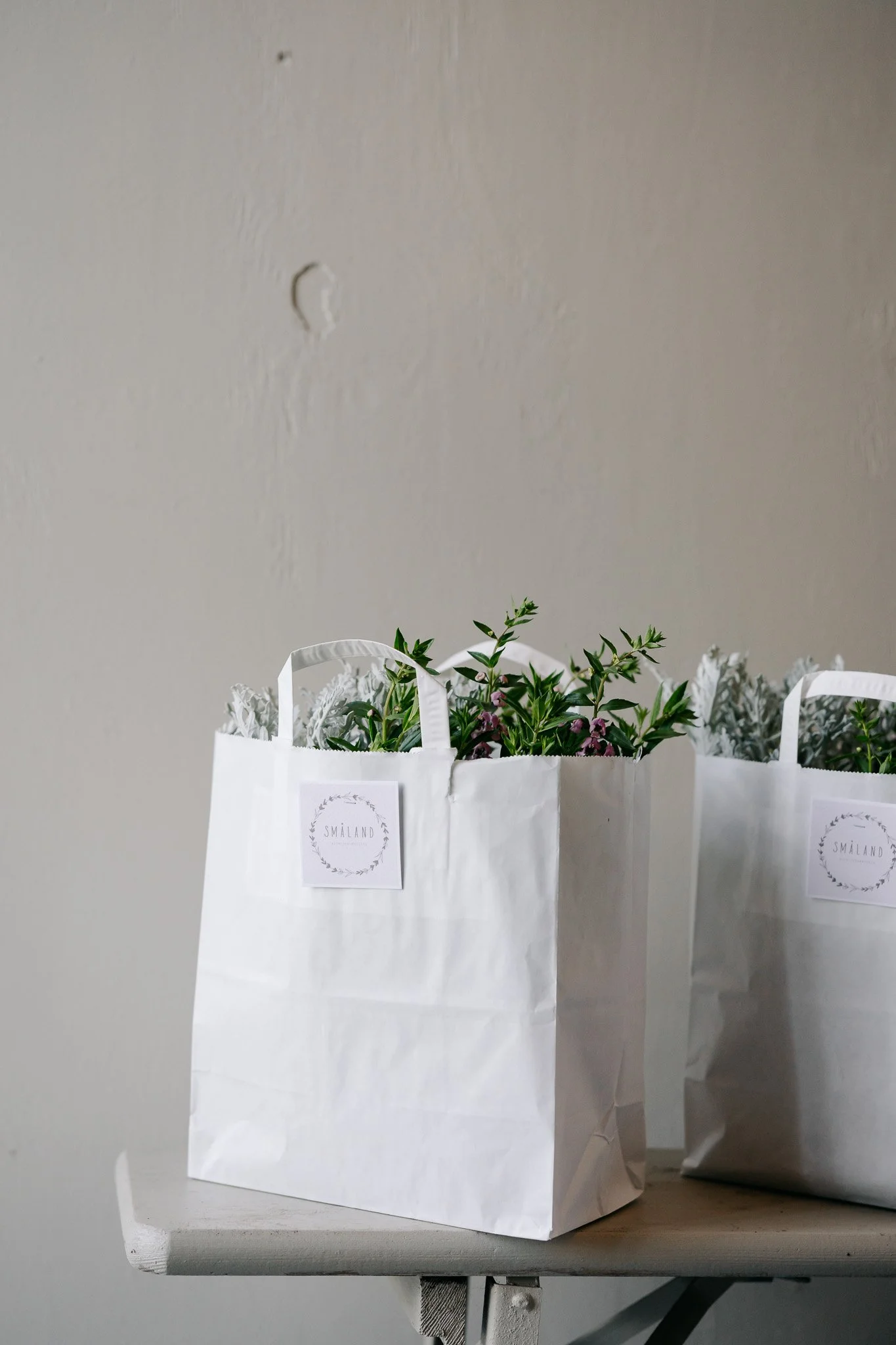 Tre hvide papirposer med grønne planter og blomster. Poserne har etiketter med teksten 'SMÅLAND' og et græsklædt blomsterdesign. Poserne står på en træbord, mod en neutral væg.
