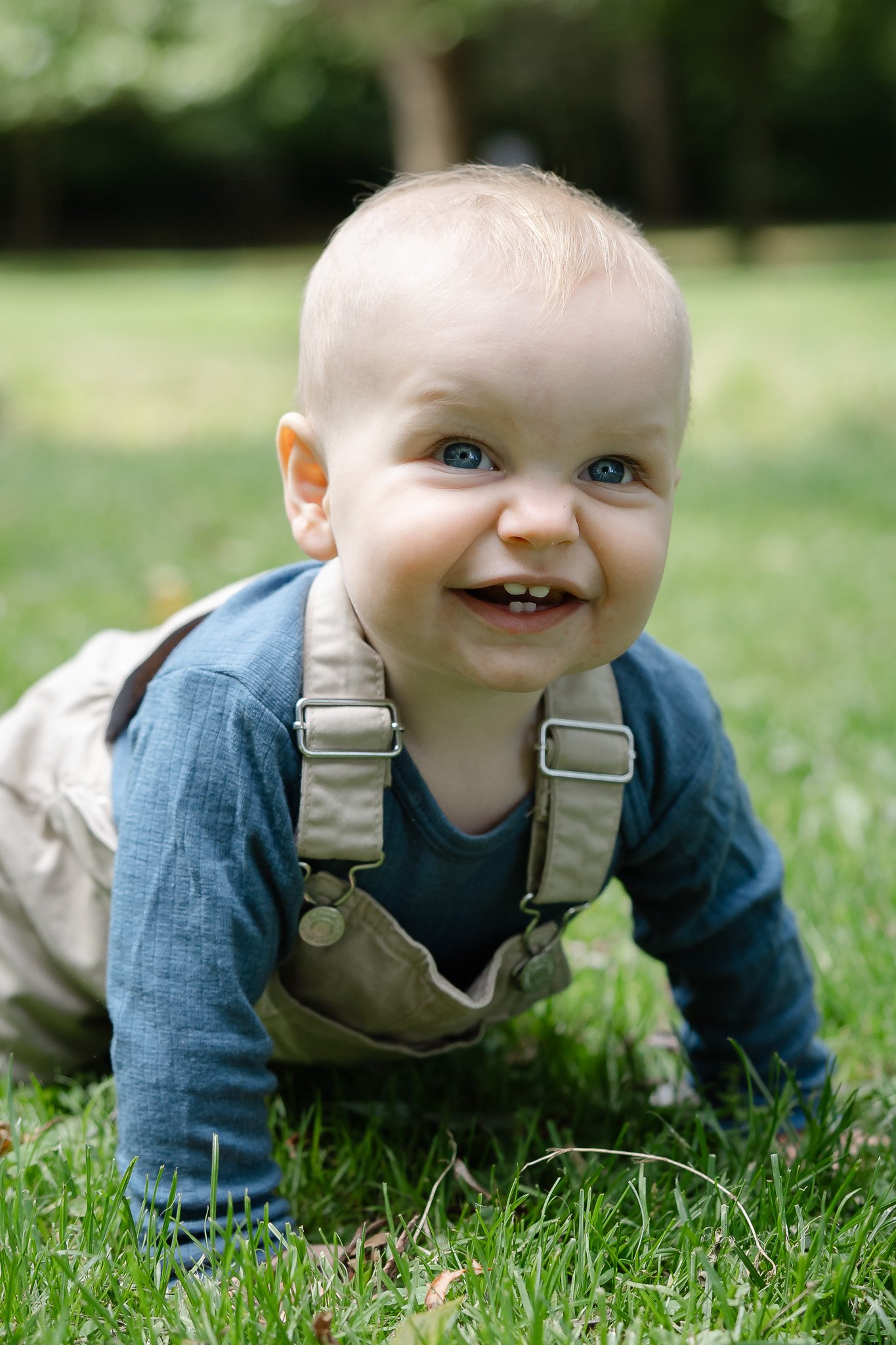 Smilende baby i blåt tøj og beige overalls, der kravler på græsset under en naturlig børnefotografering