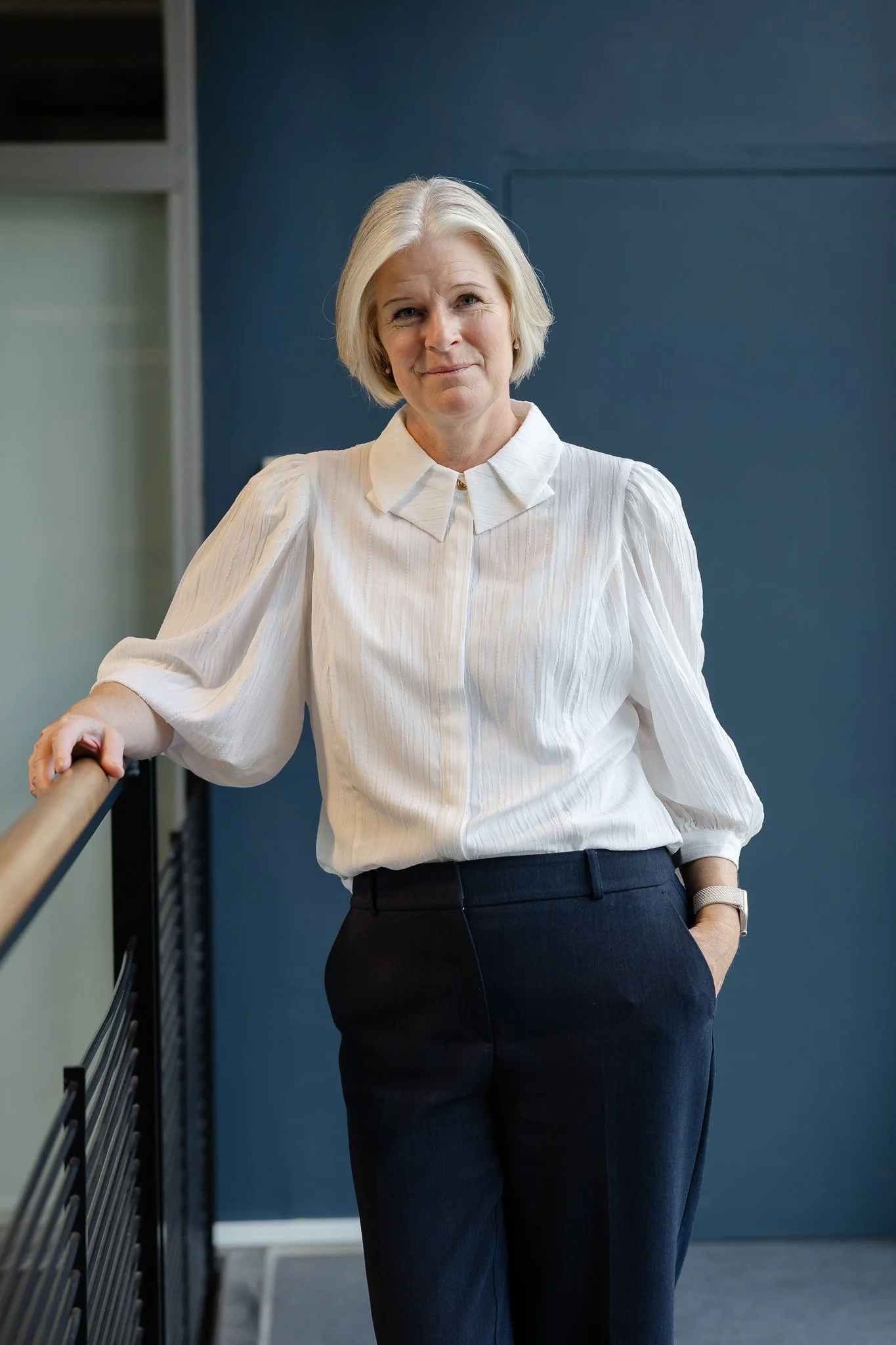 En dame med blond kort hår iført hvid skjorte og sorte bukser står ved en håndliste, med en blå væg bag sig.