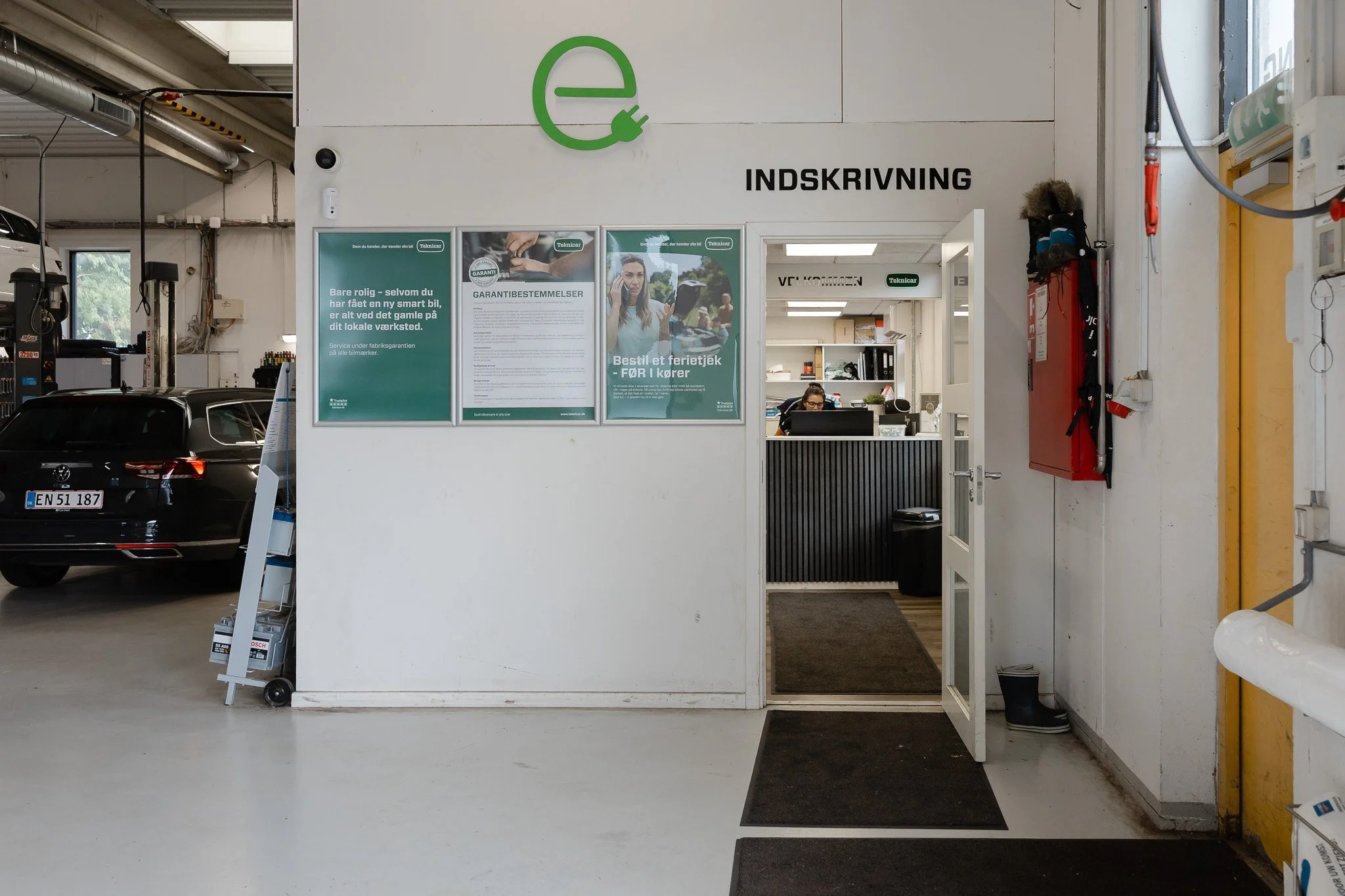 Indgang til et værksted med skiltet 'INDSKRIVNING' over døren, der fører til et skrive- og administrationsområde med en person, der arbejder ved en computer.