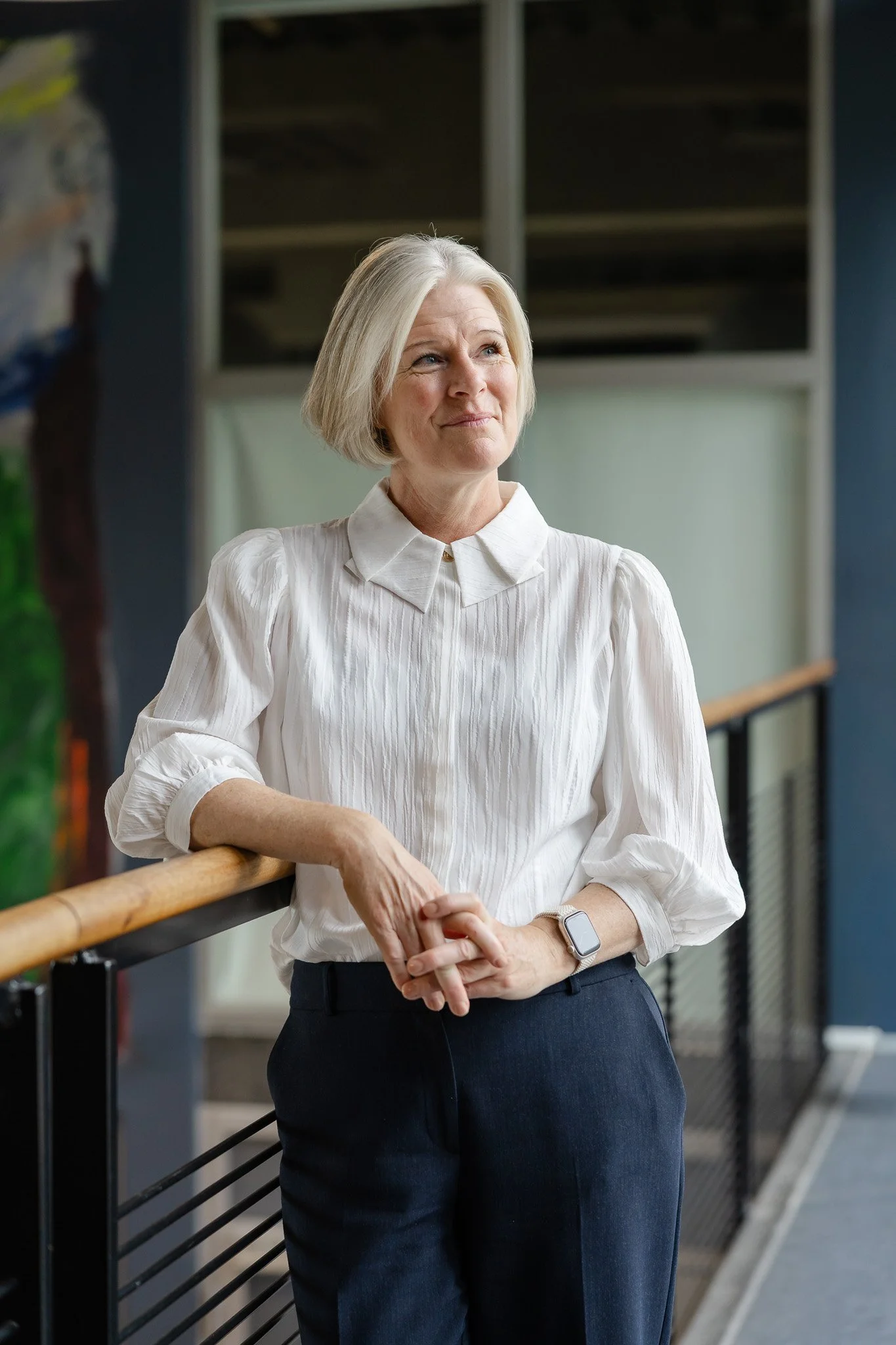 En kvinde med blond, skulderlangt hår iført hvid skjorte og mørke bukser stående ved en gelænder, smilende og kigger til siden. Baggrund med vinduer og en korridor.