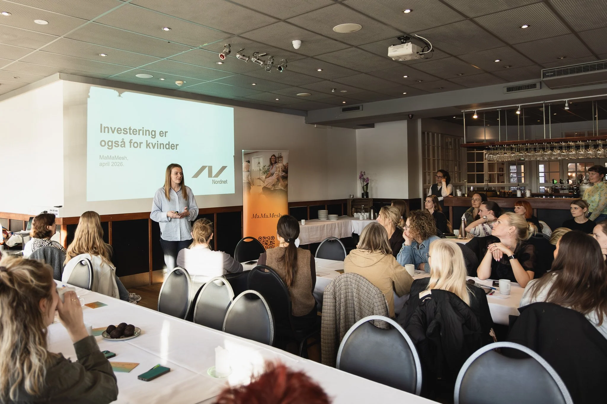 En kvinde holder en tale foran en gruppe mennesker i et konferencerum, hvor en præsentation vises på en stor skærm med teksten 'Investering er også for kvinder'.