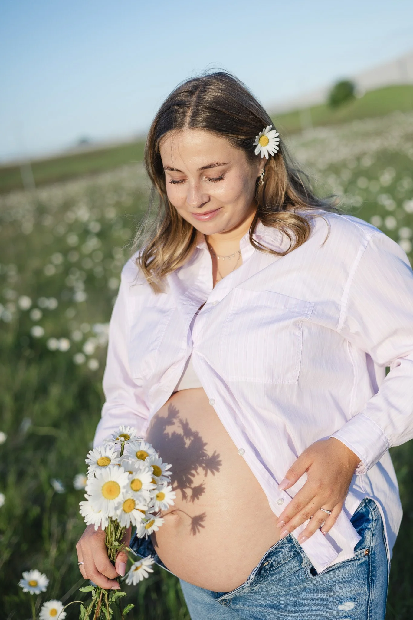 Gravid kvinde holder en buket hvide margueritter ved sin mave i et naturligt graviditetsportræt.