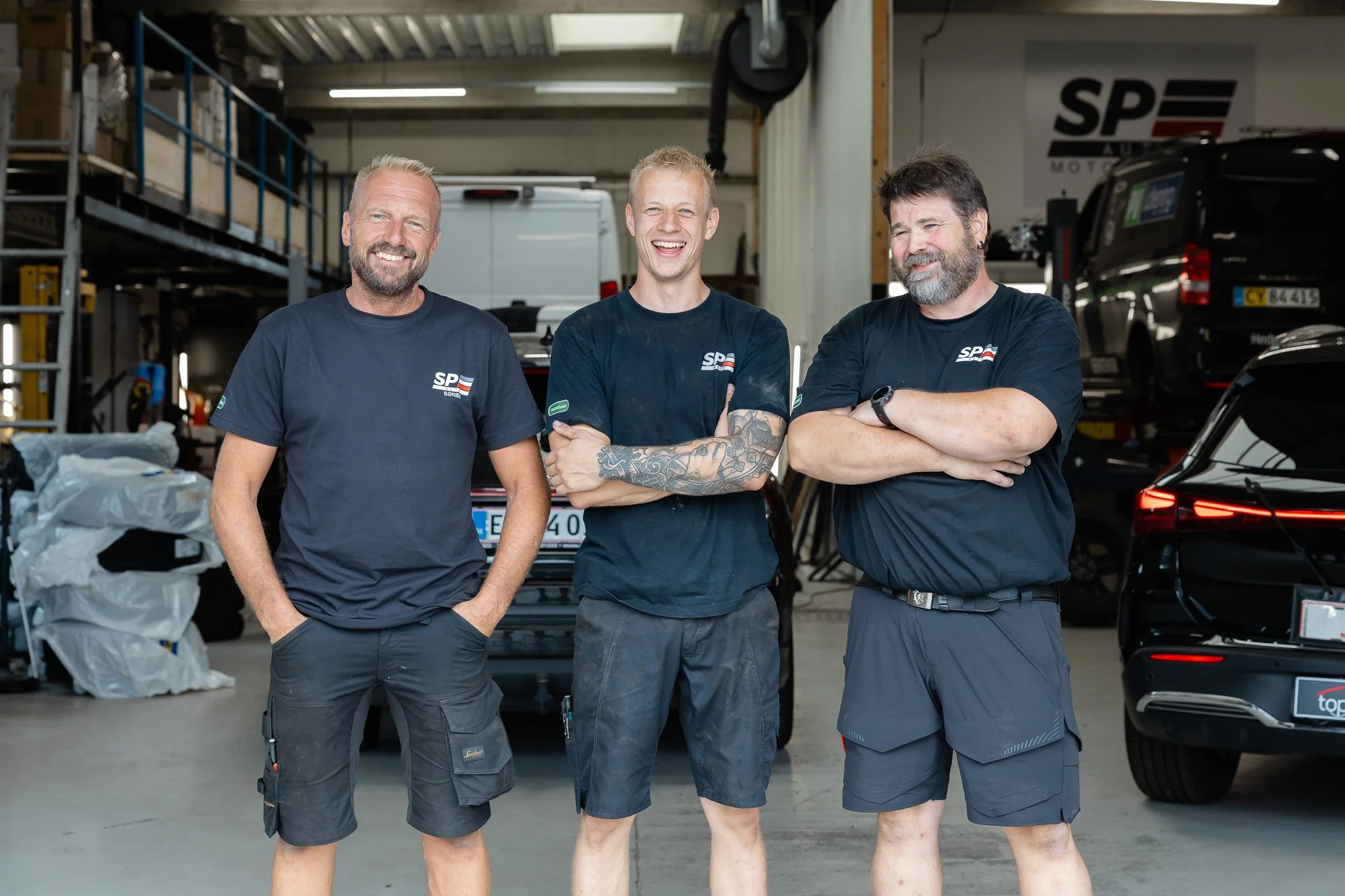 Tre mænd står sammen i en automekanikerværksted, alle iført sorte t-shirts med logo. De smiler og har armene korslagt. I baggrunden er der biler, opbevaringshylder og et værktøj.