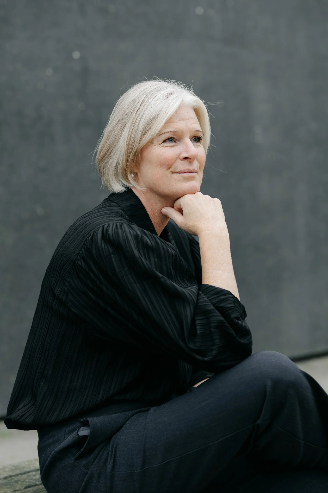 En ældre kvinde med gråt hår sidder med hagen hvilende i hånden, iført sort jakke og mørke bukser, foran en grå væg.