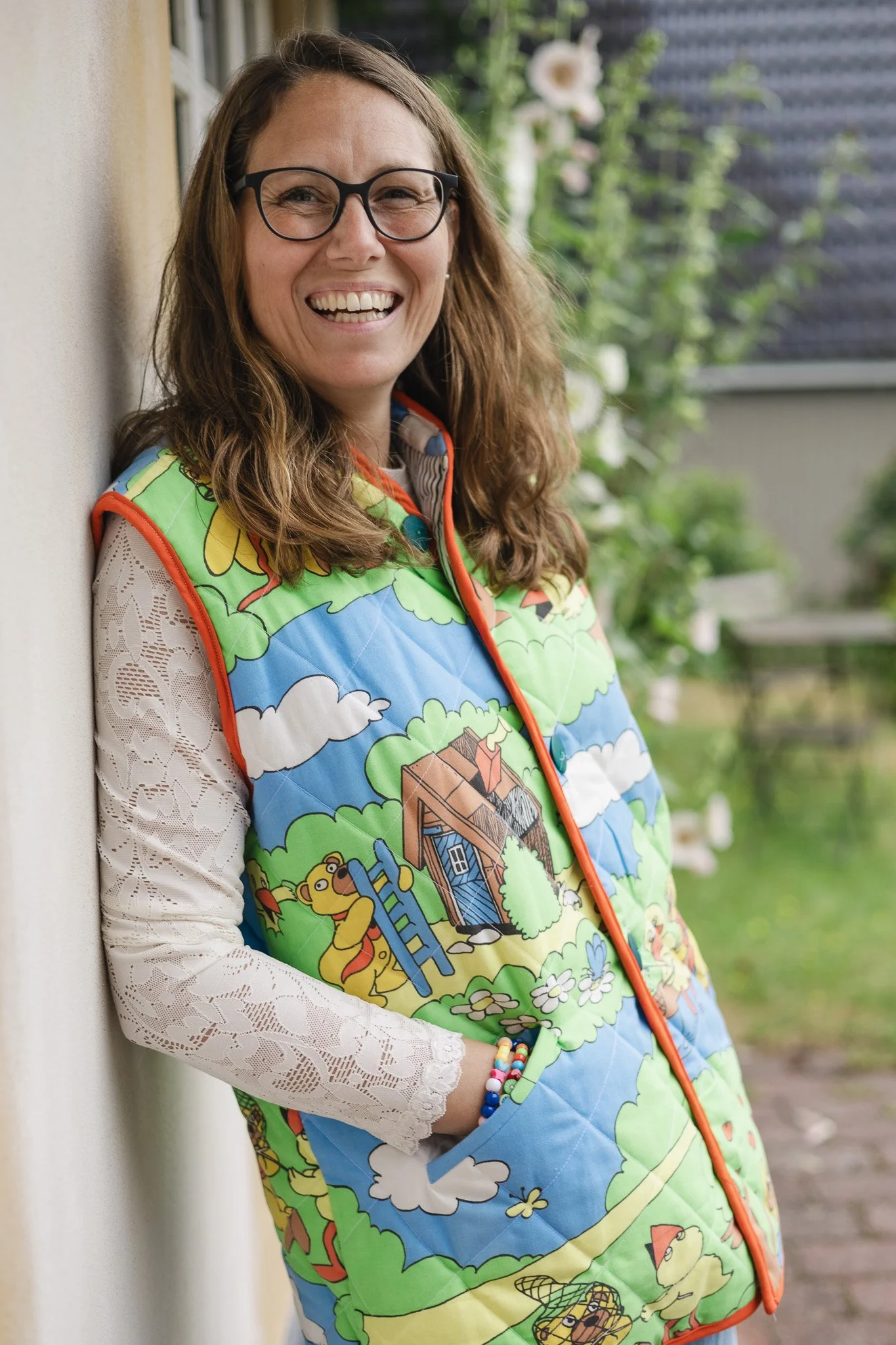 Et smilende kvindemenneske med briller, iført en farverig vest med tegneserie-illustrationer, stående udendørs ved en mur med vækst i baggrunden.