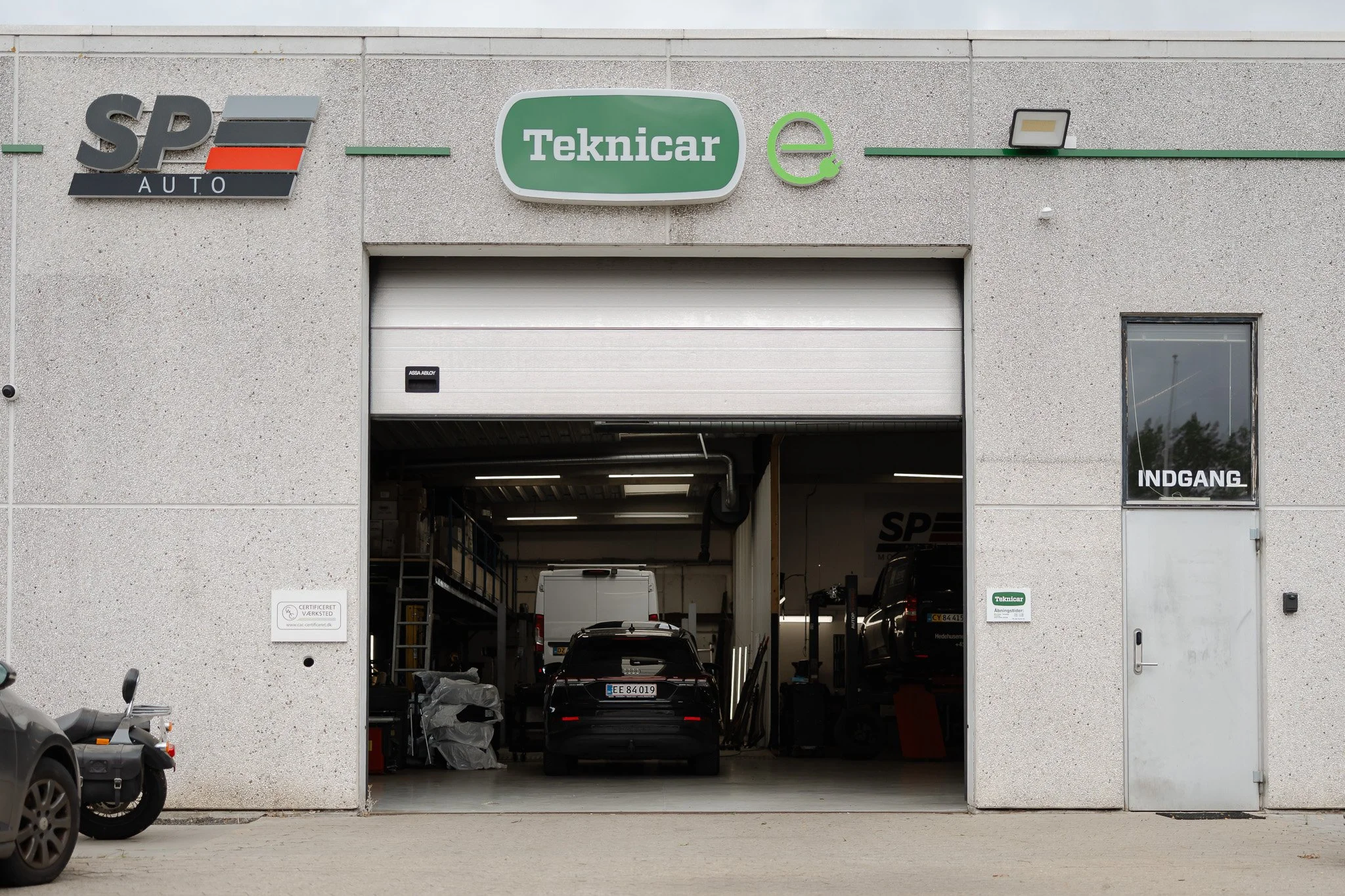 Bilsalgs- og værkstedshal med åbent garageport, flere biler parkeret indendørs, skilte med logoerne 'SPE AUTO' og 'Teknicar', og en elektrisk opladningssymbol over døren.