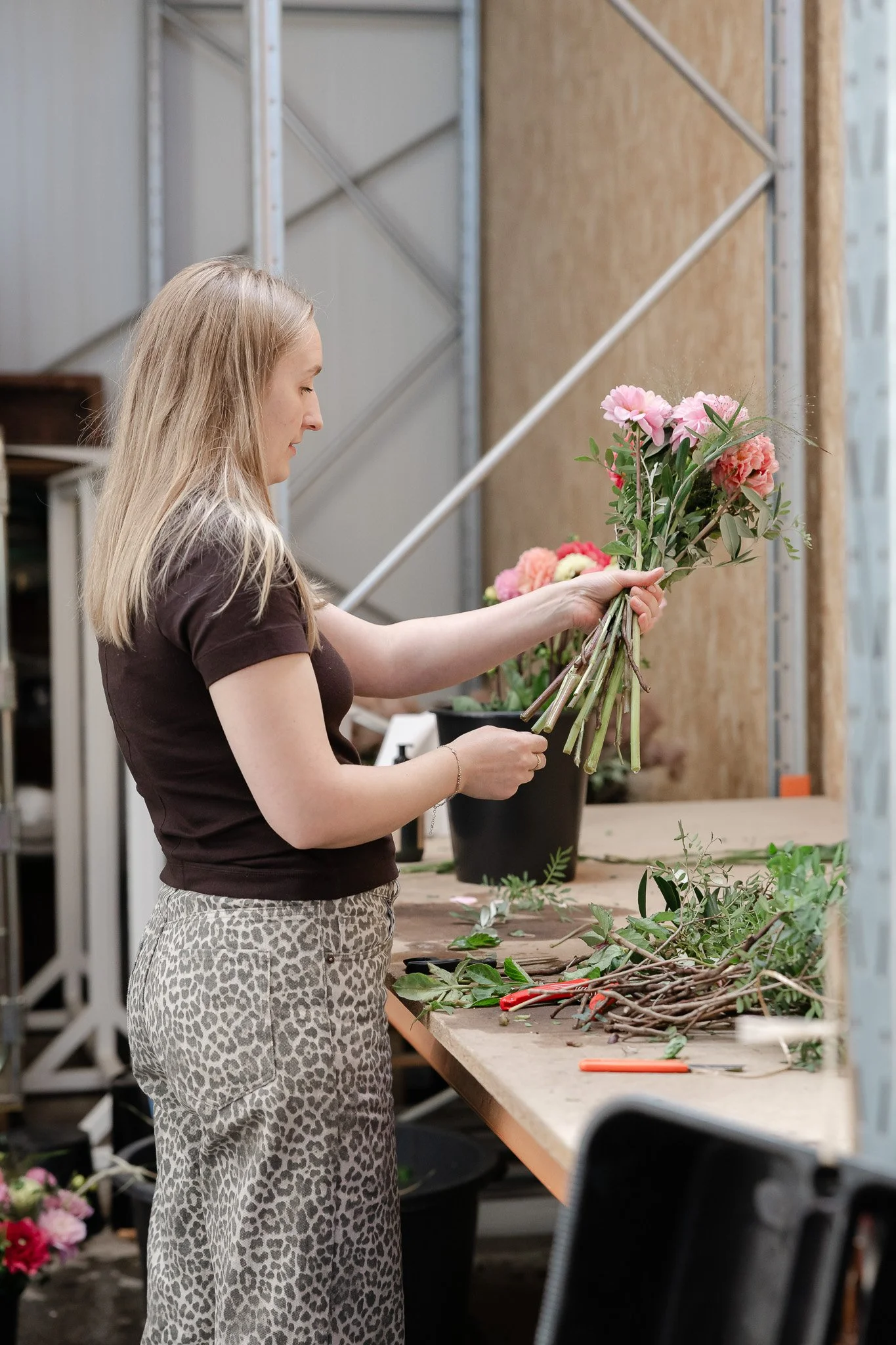 Kvinde med blond hår, iført leopardprint bukser og sort t-shirt, arbejder med blomster på et bord i et værksted eller blomsterbutik.