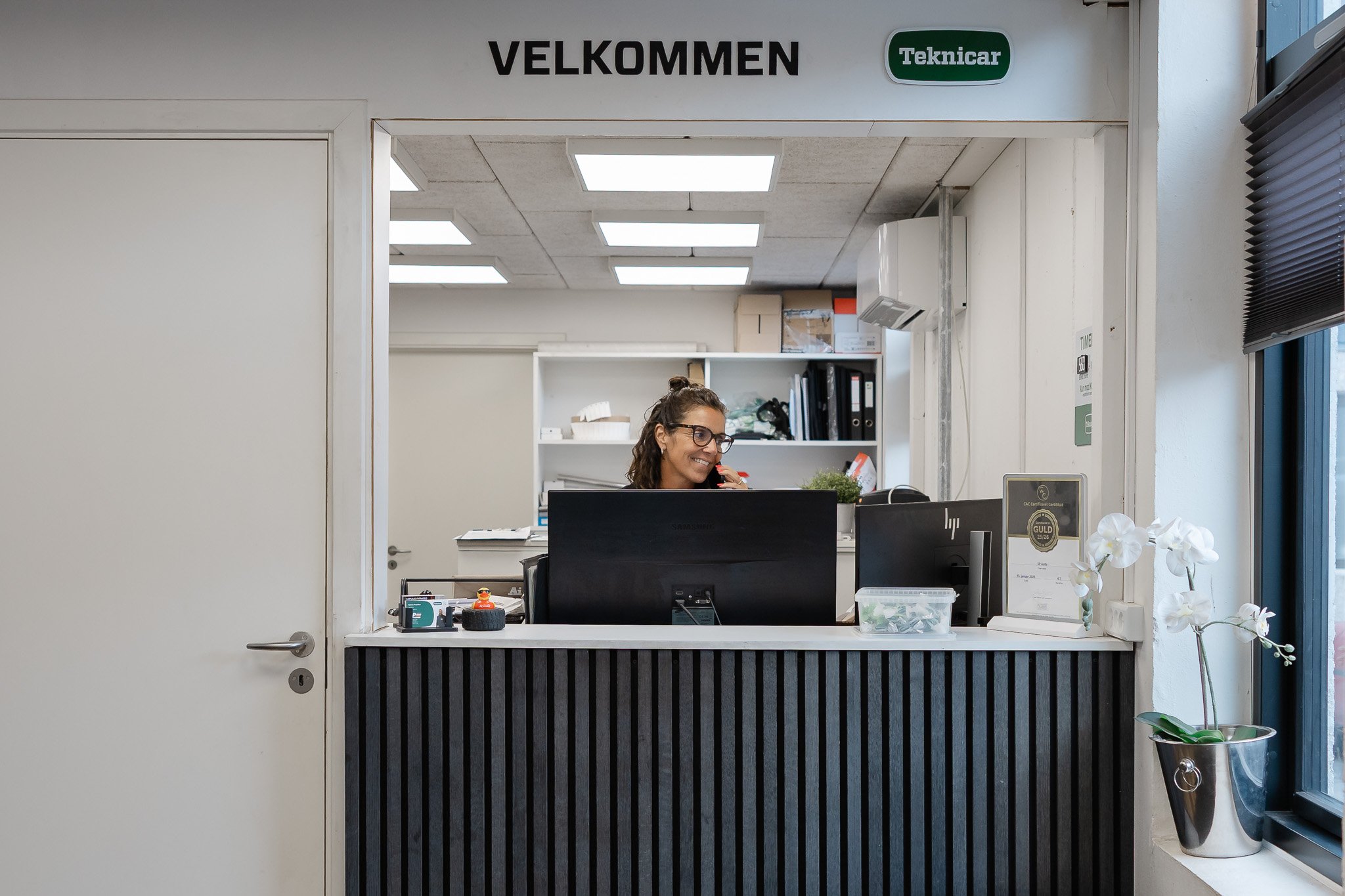 Reception i en bilforretning med en smilende kvinde, computer, planter og skilte med 'Velkommen' og 'Teknicar'
