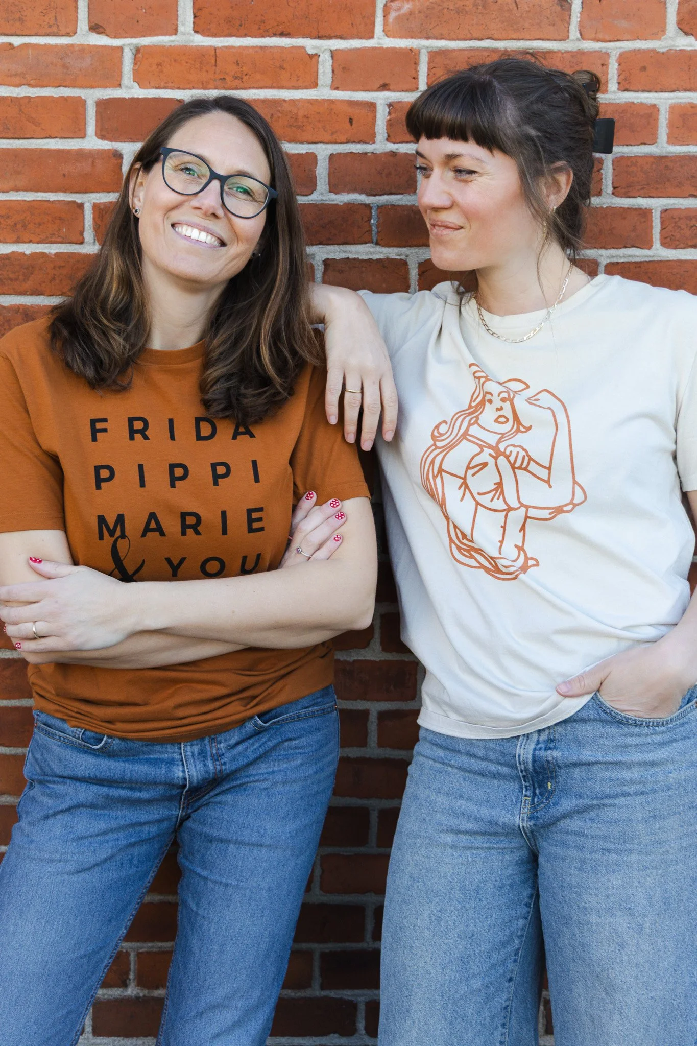 To kvinder står sammen mod en rød mur. Den venstre kvinde har brunt hår, briller og en orange T-shirt med teksten 'FRIDA PIPPI MARIE & YOU'. Den højre kvinde har brunt hår med pandehår, og hun har en lys T-shirt med en figur af en kvinde, der flexer 