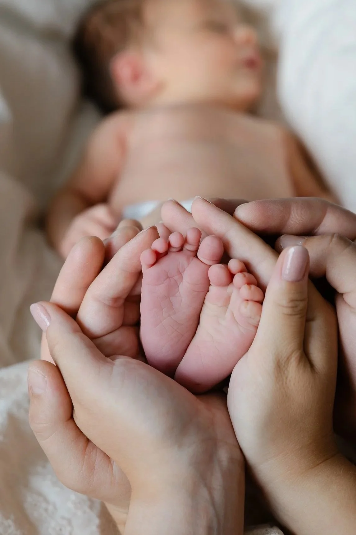 Billede af en voksen, der holder et nyfødt barns fødder i hænderne, med en sovende baby i baggrunden.