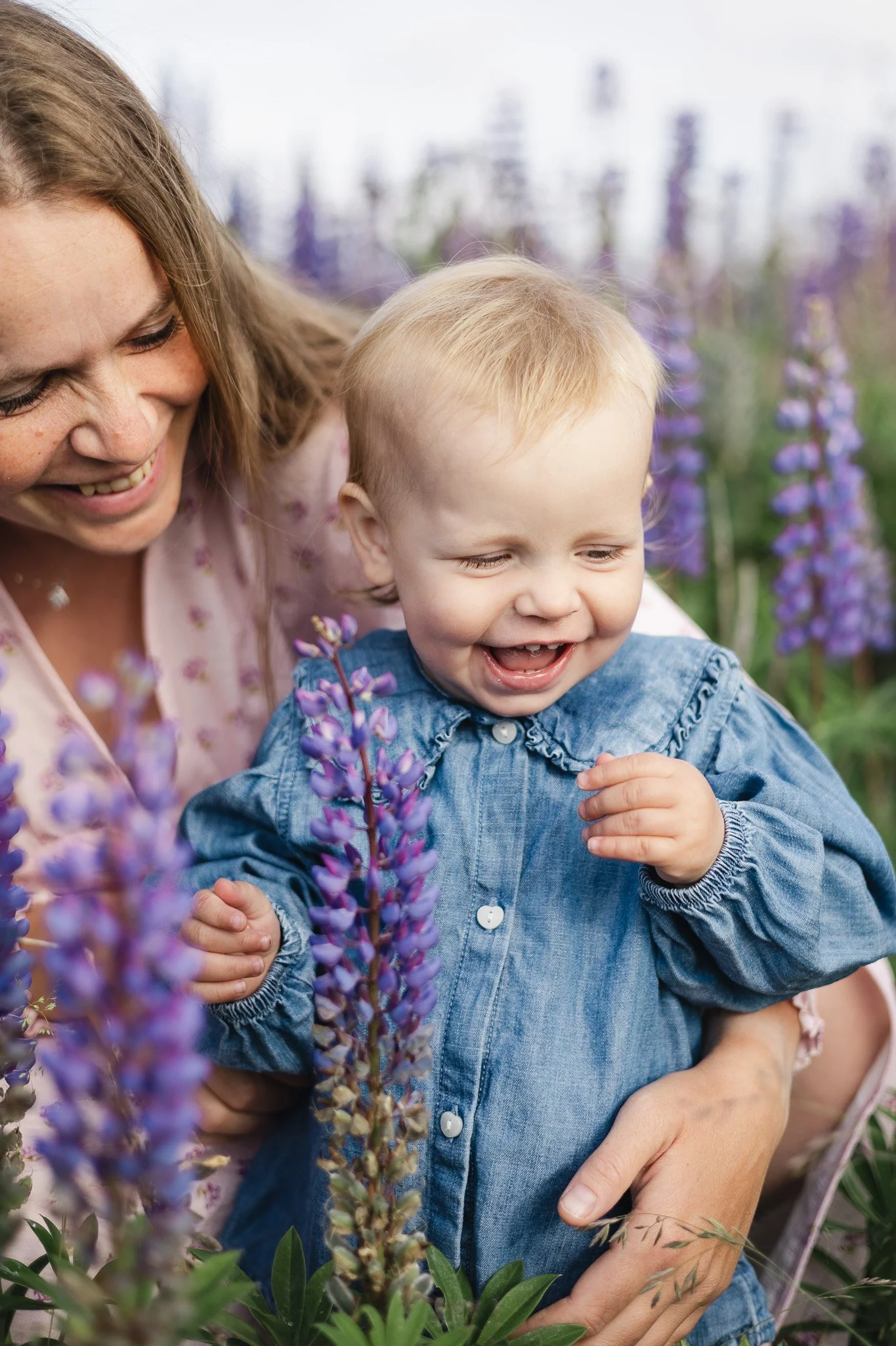 En glad voksen og et barn i et blomsterbed med purple blomster og grønne blade, smilende og nyder tiden sammen.
