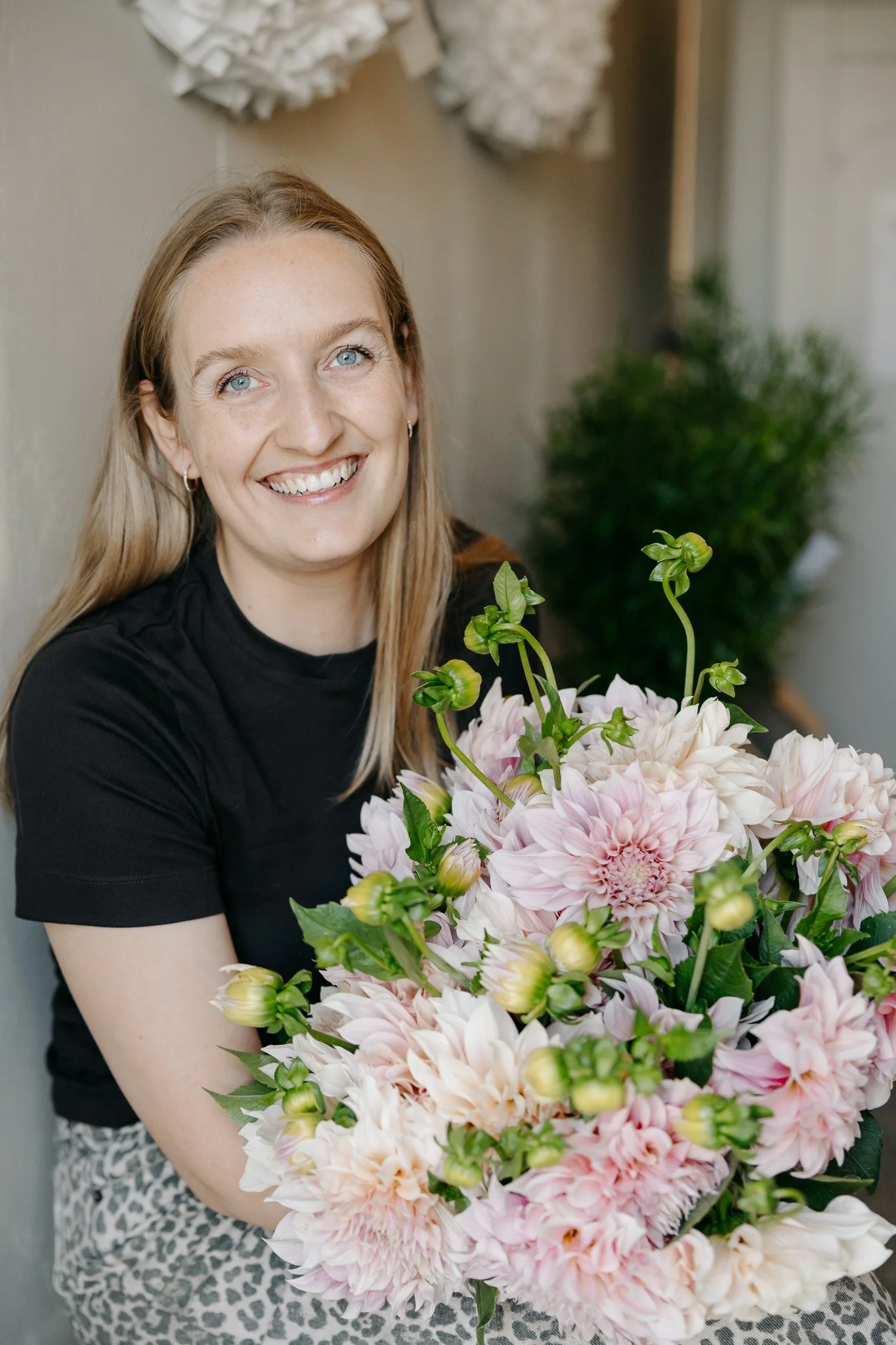 En glad kvinde med blond hår og blå øjne smiler mens hun holder en stor buket lyserøde og hvide blomster. Hun sidder foran en beige væg med dekorative hvide papirkugler.
