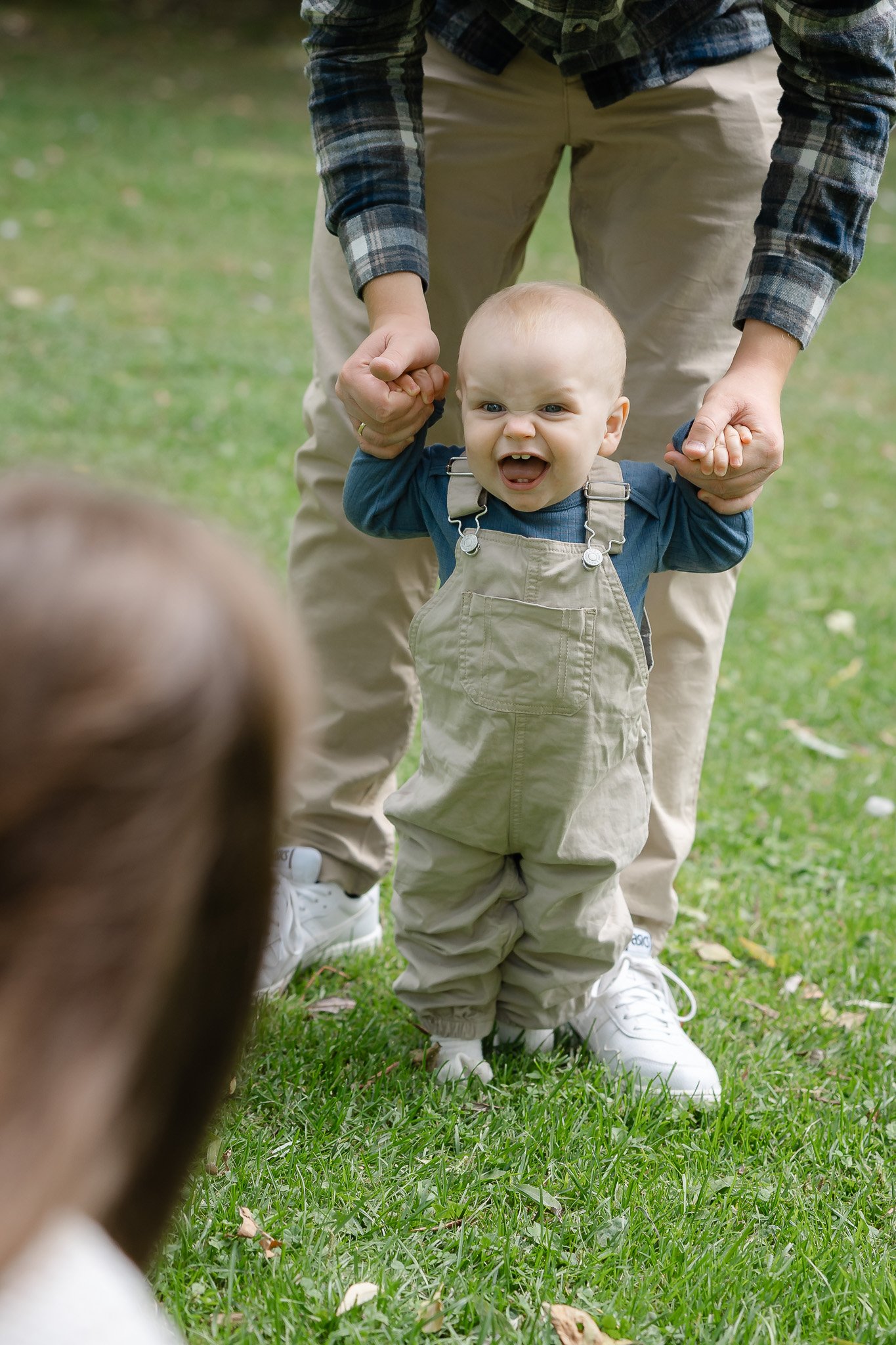 Smilende baby tager sine første skridt med støtte fra sine forældre under en udendørs familiefotografering.