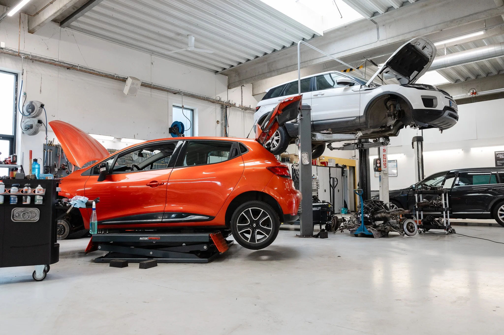 En bilværksted med to biler, hvor den ene er en orange sportscar og den anden er en hvid bil, der er løftet op på en ophængningsramme til reparation. Der er værktøjer og maskiner omkring, og at det er en industrielt indrettet garage.