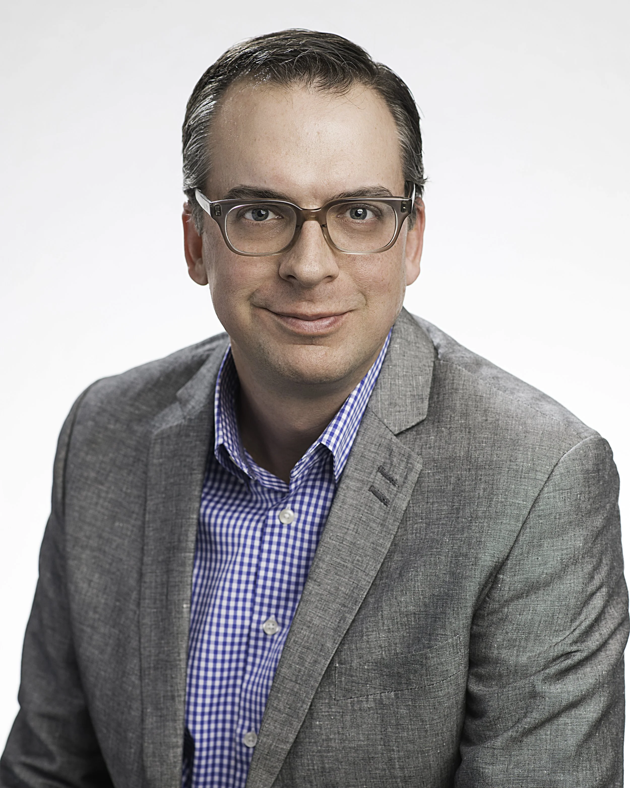 Photo of Chris White glasses, a blue shirt, and a gray sports coat in front of a white background